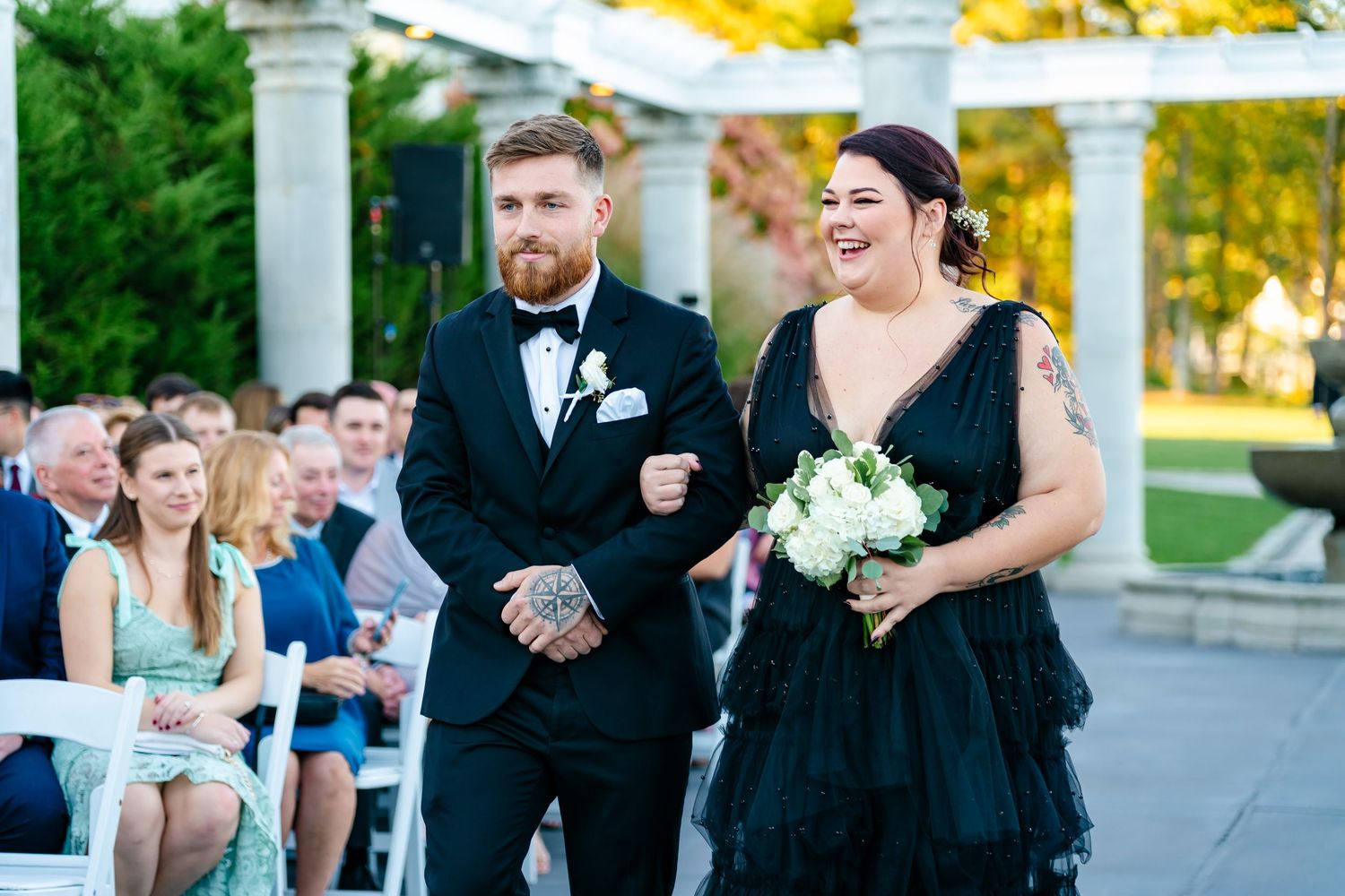 Wedding ceremony guests seated outdoors under white pillars and autumn foliage at an elegant garden venue.