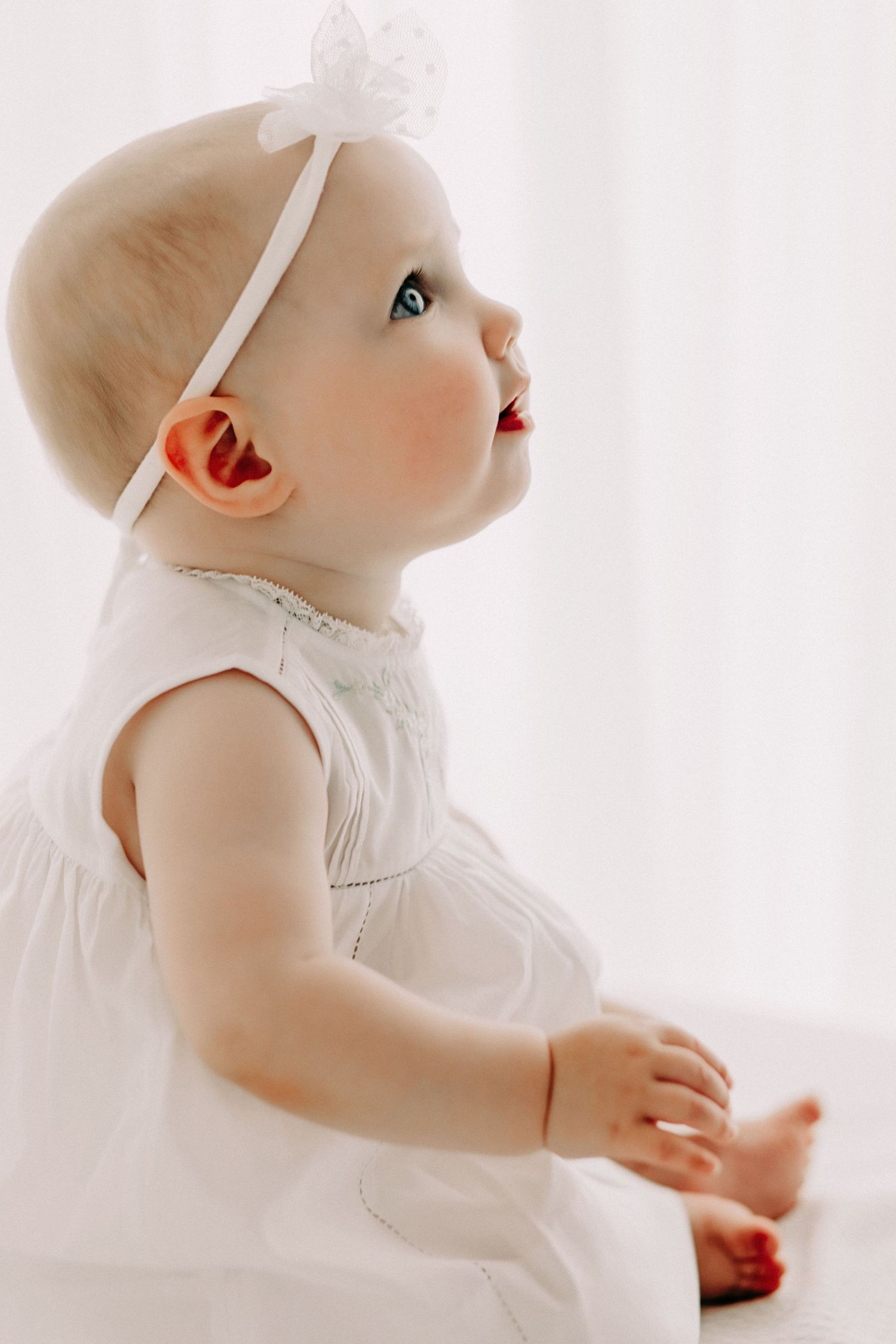 Baby in white dress and bow headband looking up with wonder.
