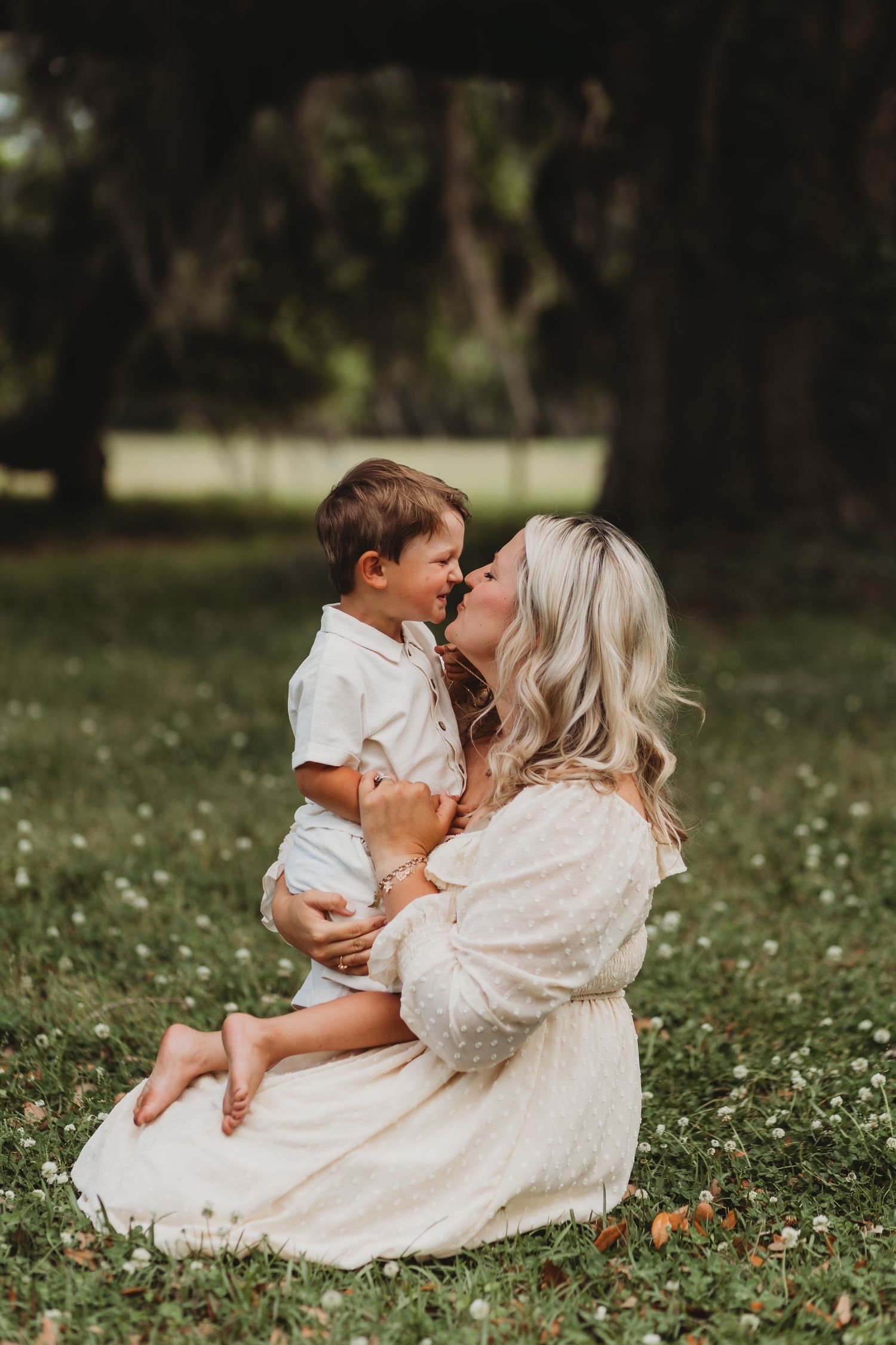 Mother in flowing white dress shares tender moment with child on flowering grass.