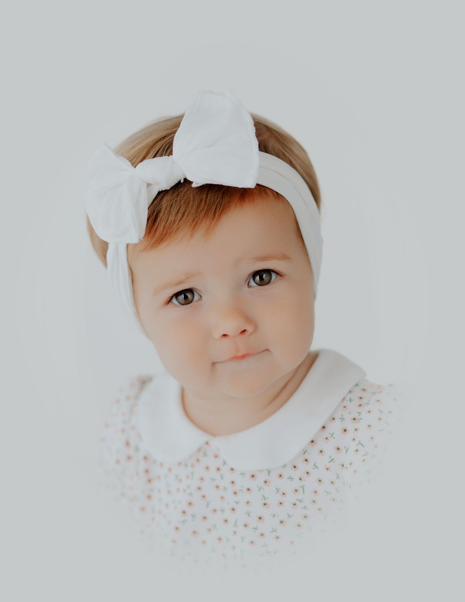 Close up portrait of baby in white headbow and polka dot dress collar against neutral backdrop.