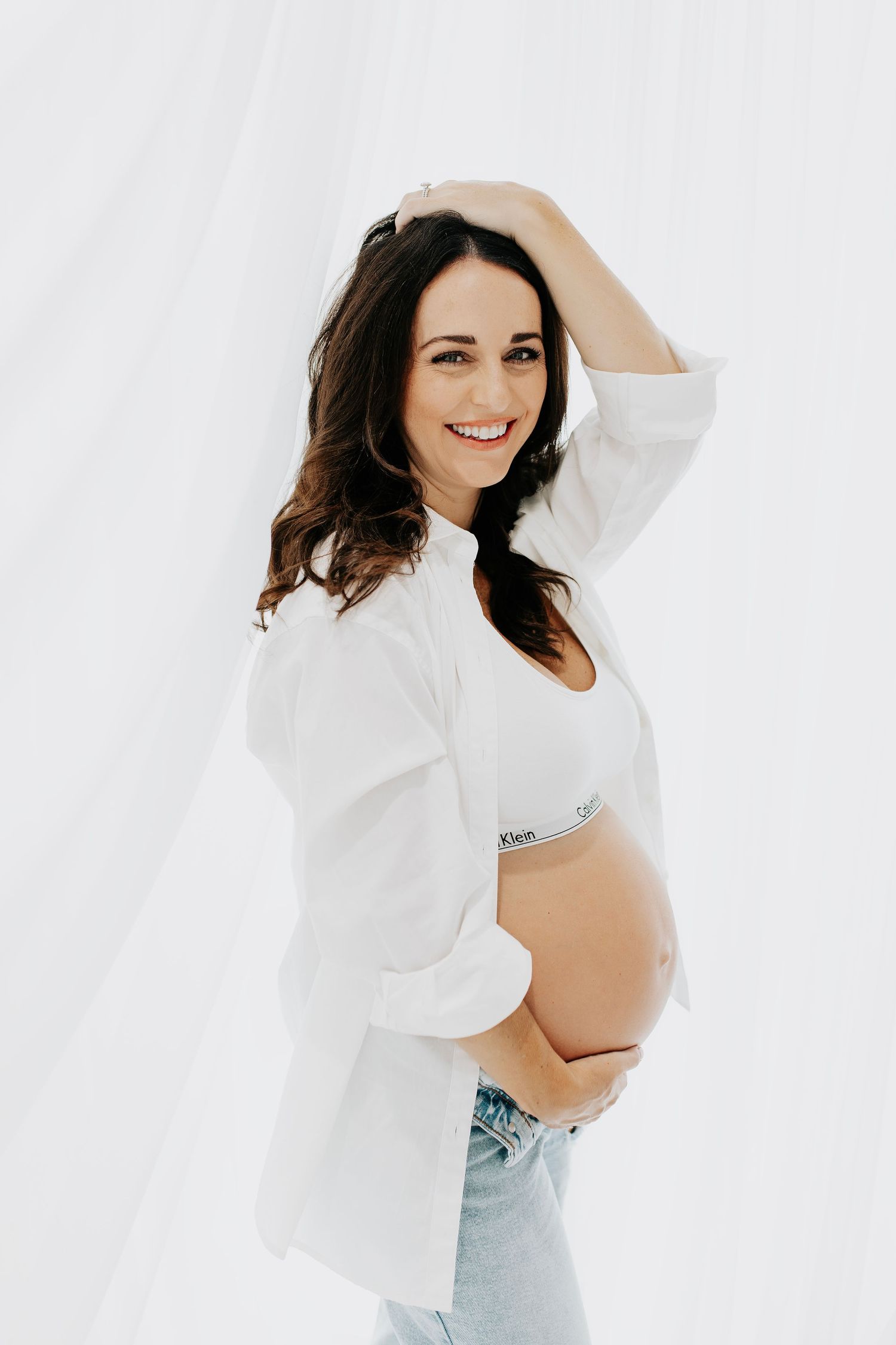 Woman in white top poses joyfully against white backdrop during maternity photoshoot.