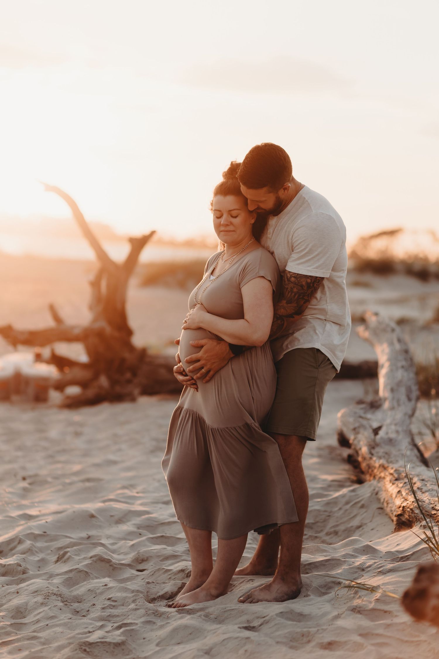 Expecting couple embraces on sandy beach during sunset maternity photography session.