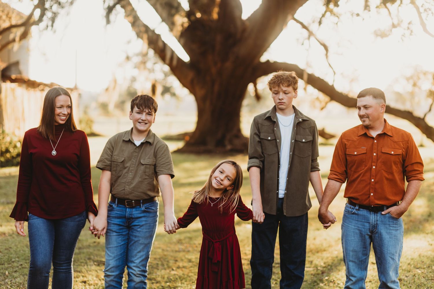 Family in coordinating burgundy and brown outfits walks together holding hands in outdoor setting.