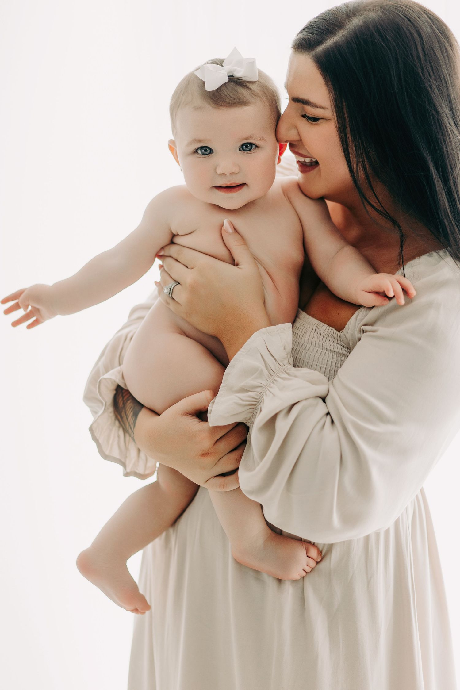 Mother in white dress holds smiling baby wearing white bow during photo session.