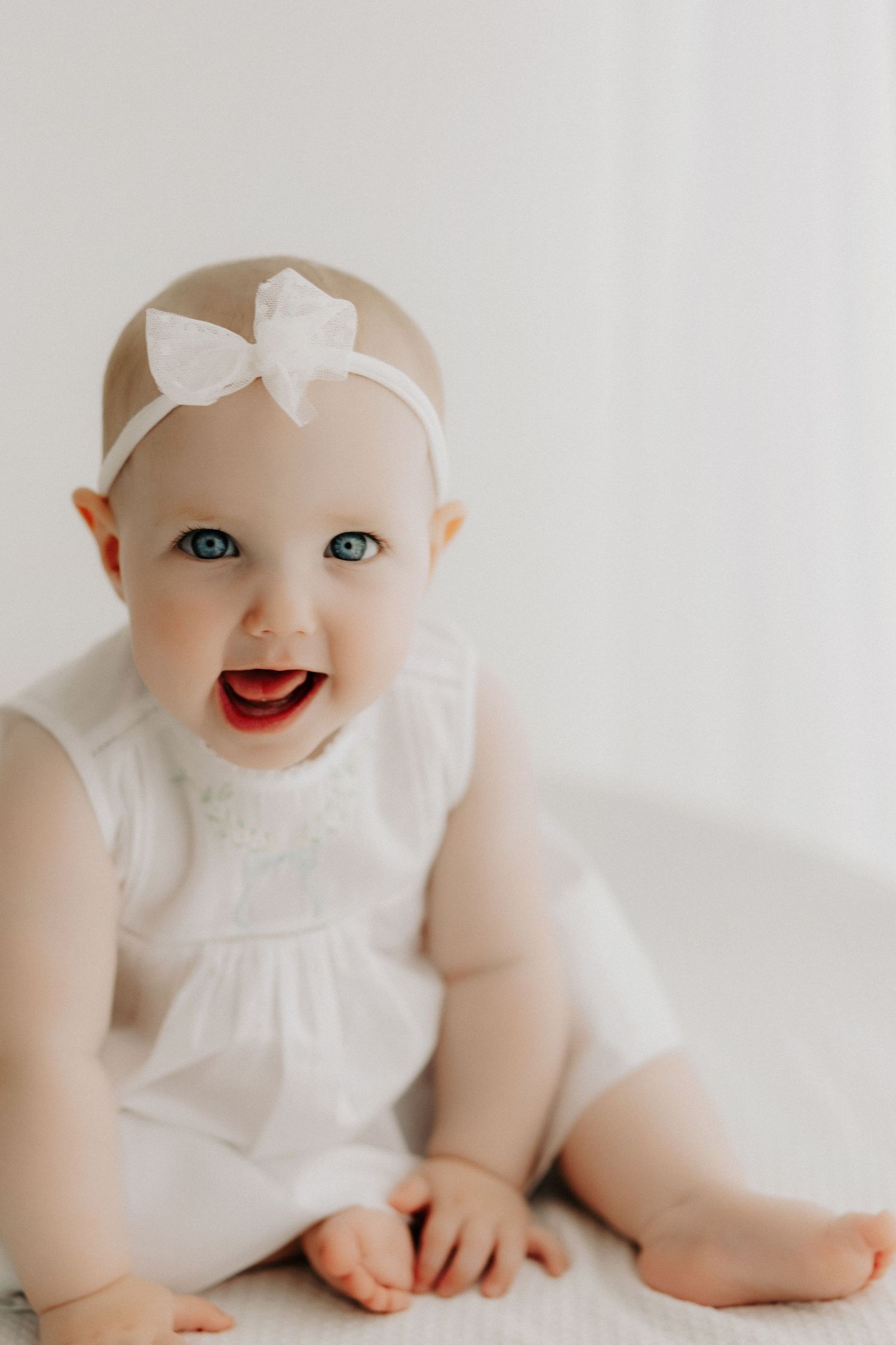 Joyful baby in white dress and bow headband smiling at camera.