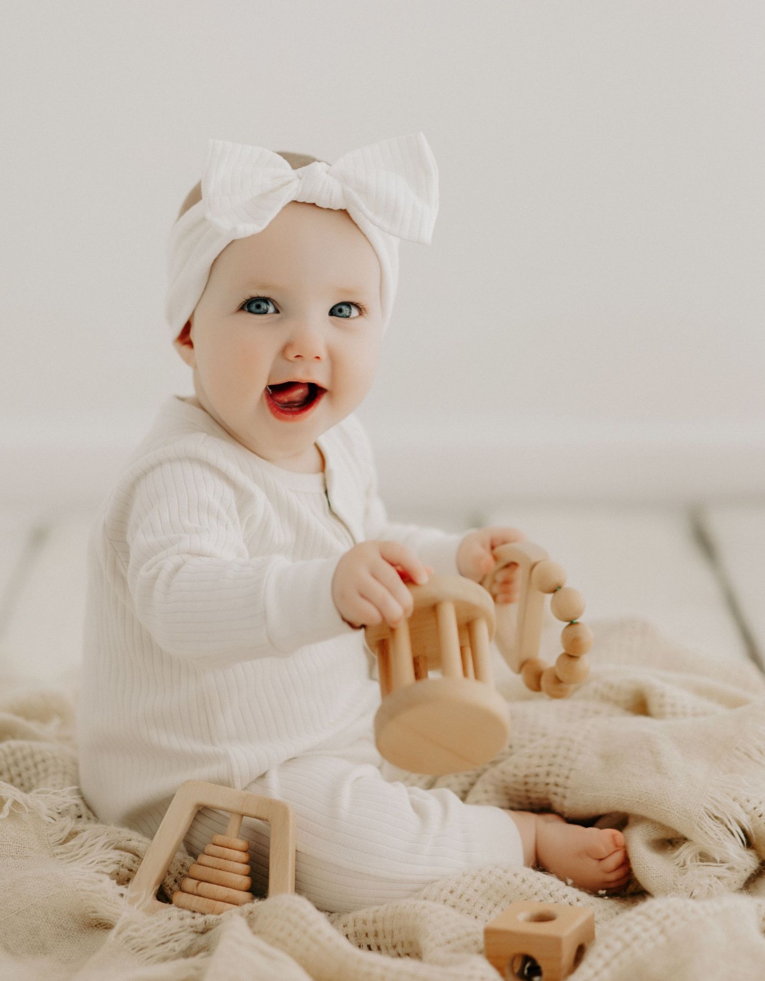 Baby in white outfit plays with wooden toys while sitting on textured blanket.
