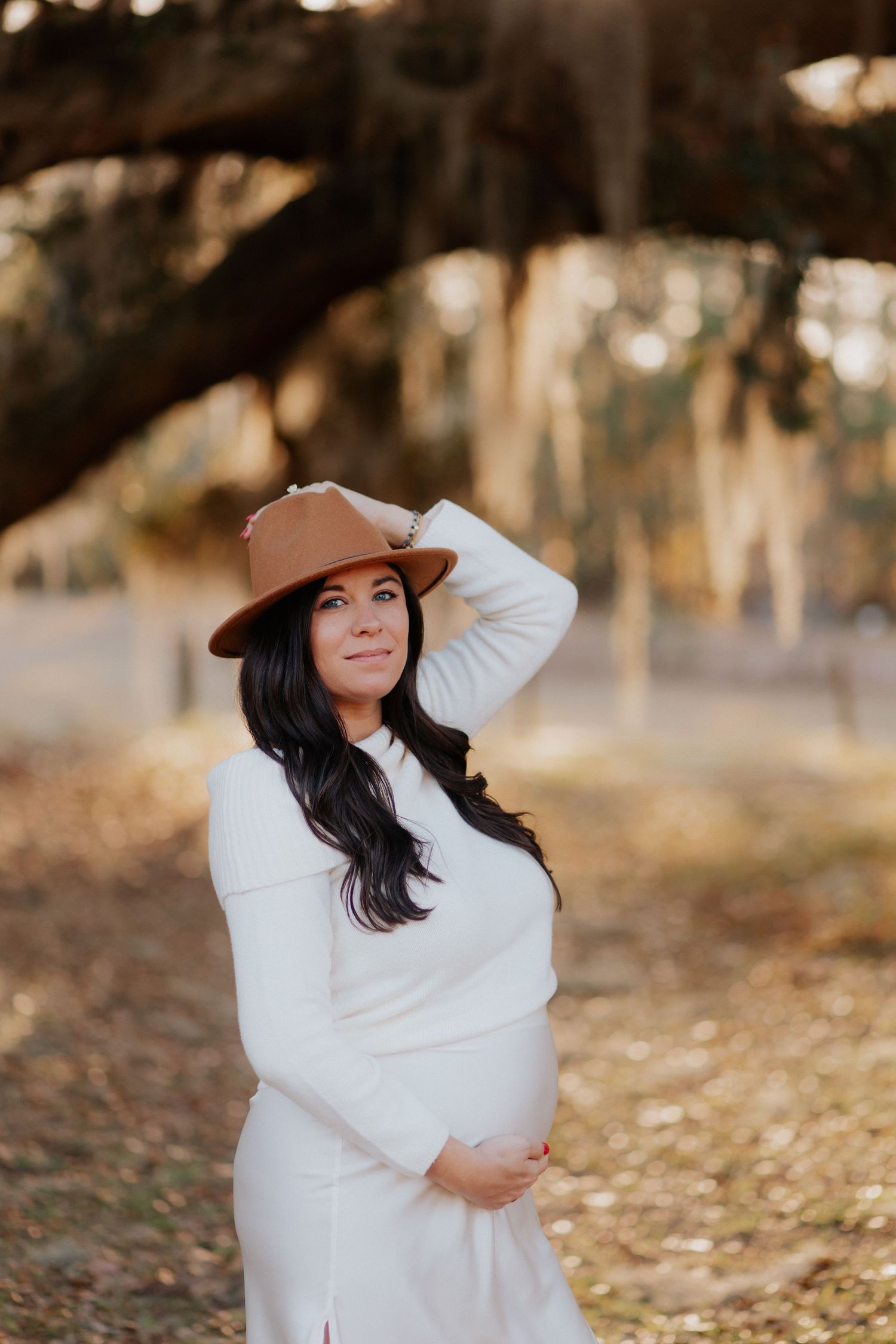 Woman in white dress poses confidently with a brown hat in a dreamy outdoor setting.