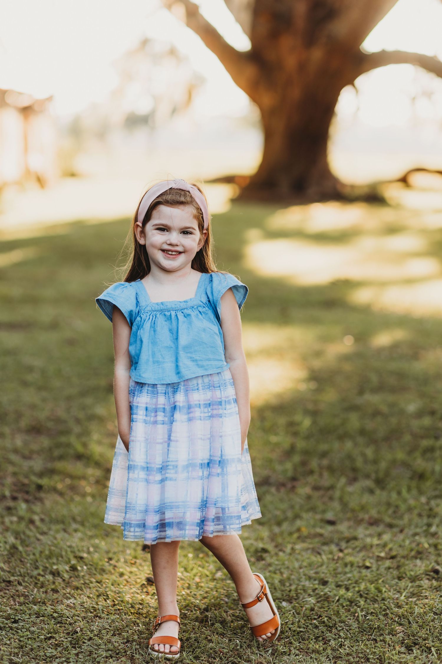 Little girl in blue tie-dye dress smiles while standing on grass during golden hour.