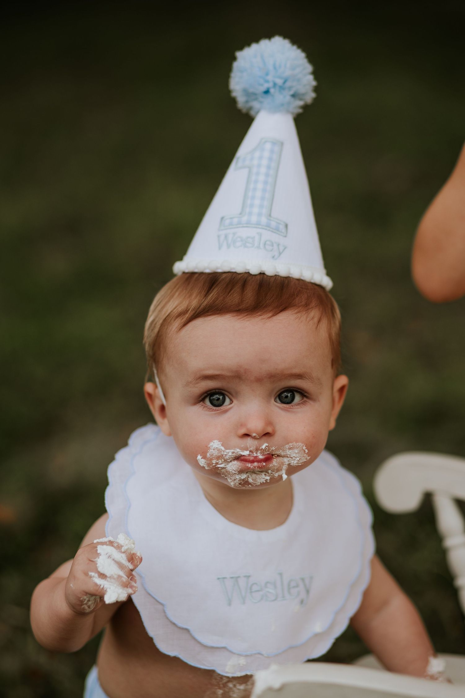 Baby in party hat and bib with cake frosting on face during first birthday celebration.