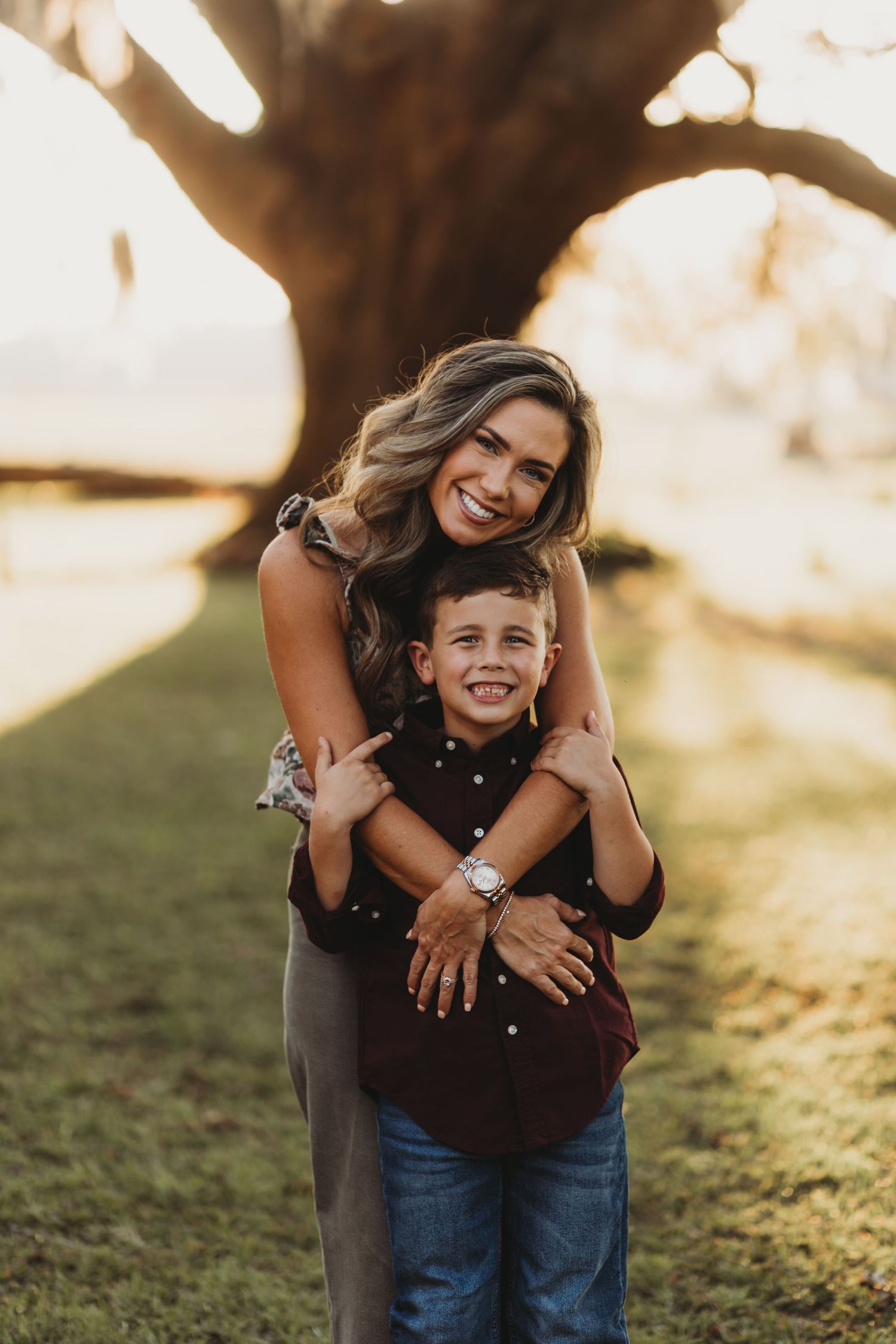 Parent and child hugging and laughing together in a sunny outdoor setting with trees in background.
