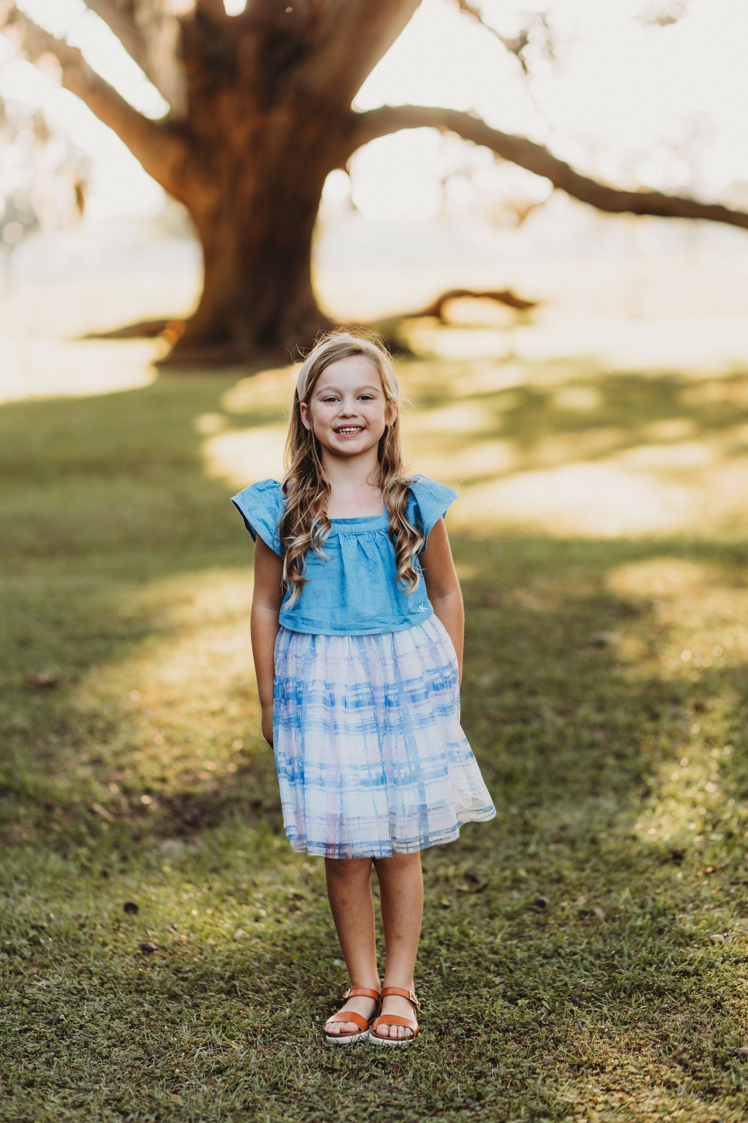 Young child in blue tie-dye dress poses happily on grass during outdoor photoshoot.