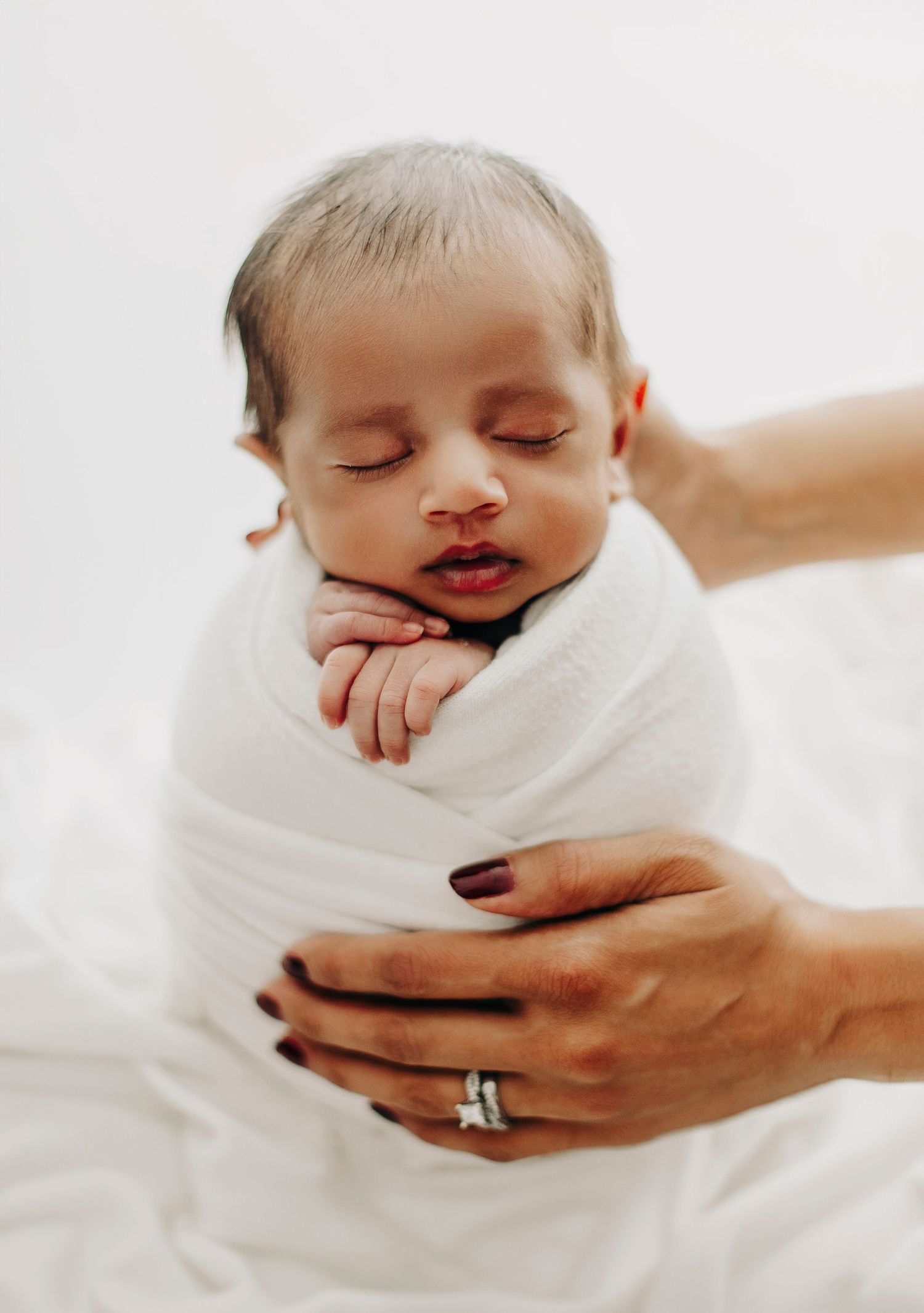Sleeping newborn wrapped in white being gently held by caring hands.