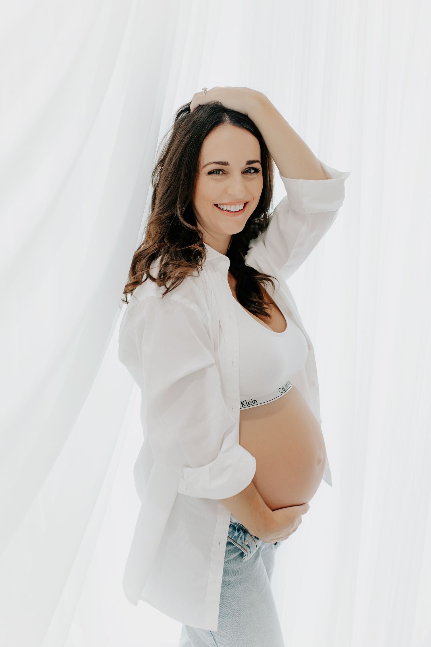 Maternity portrait of woman in white shirt and jeans against white backdrop.