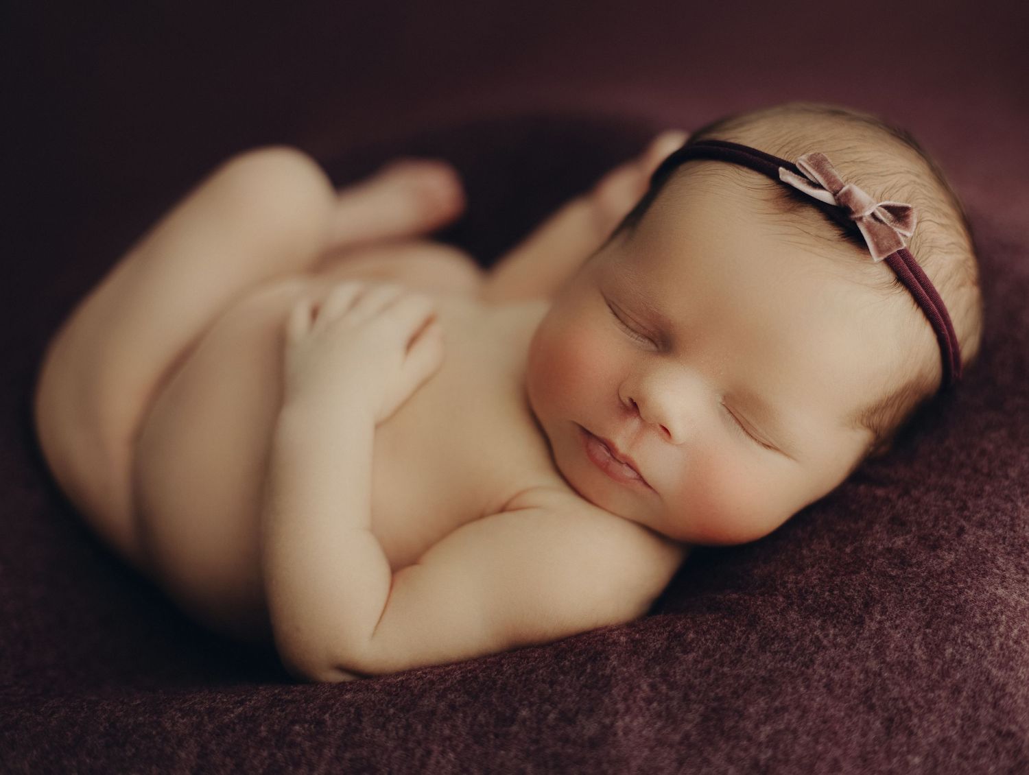 Sleeping newborn with thin headband resting on dark textured blanket.