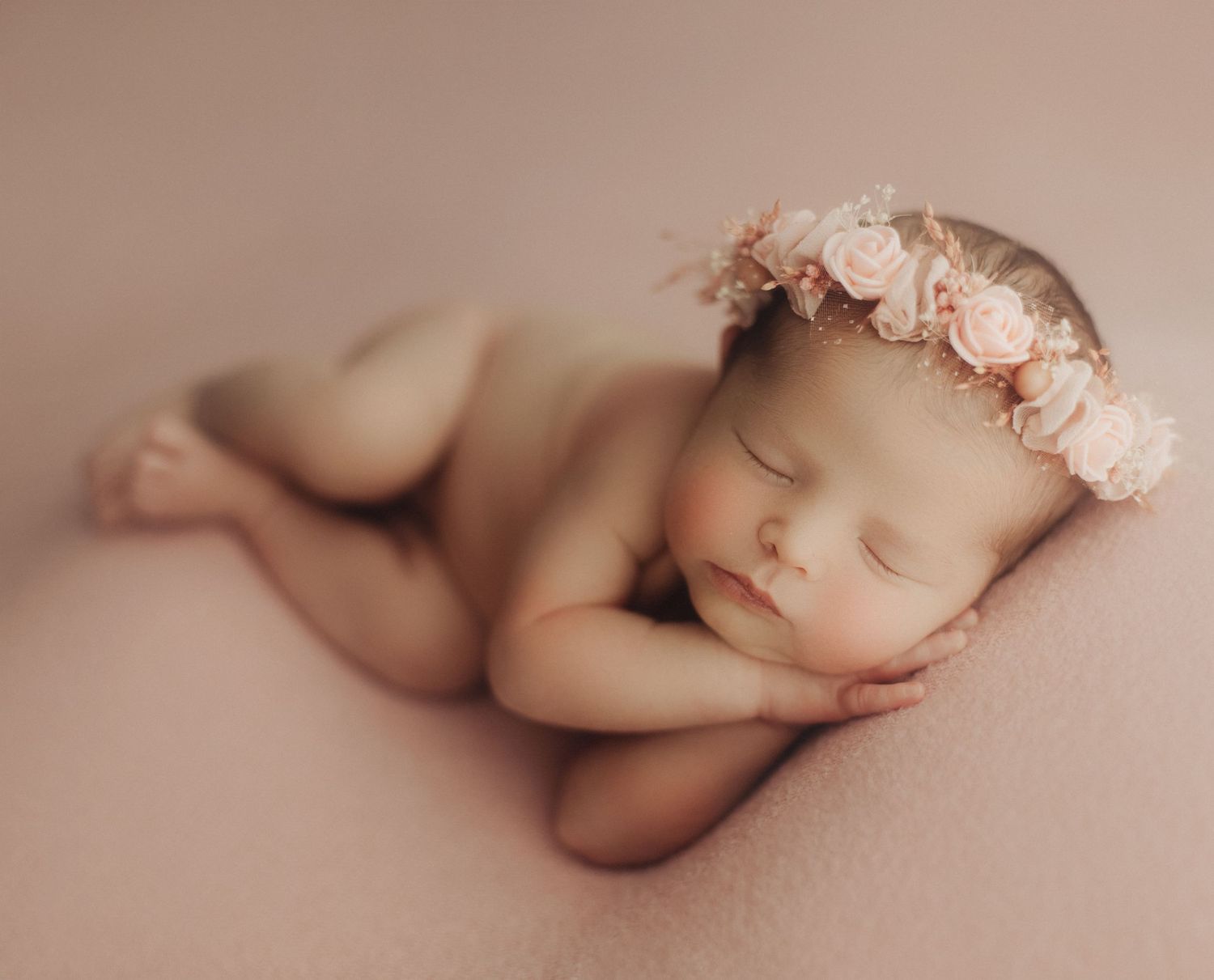 Peaceful sleeping newborn wearing delicate pink floral crown on soft beige backdrop.