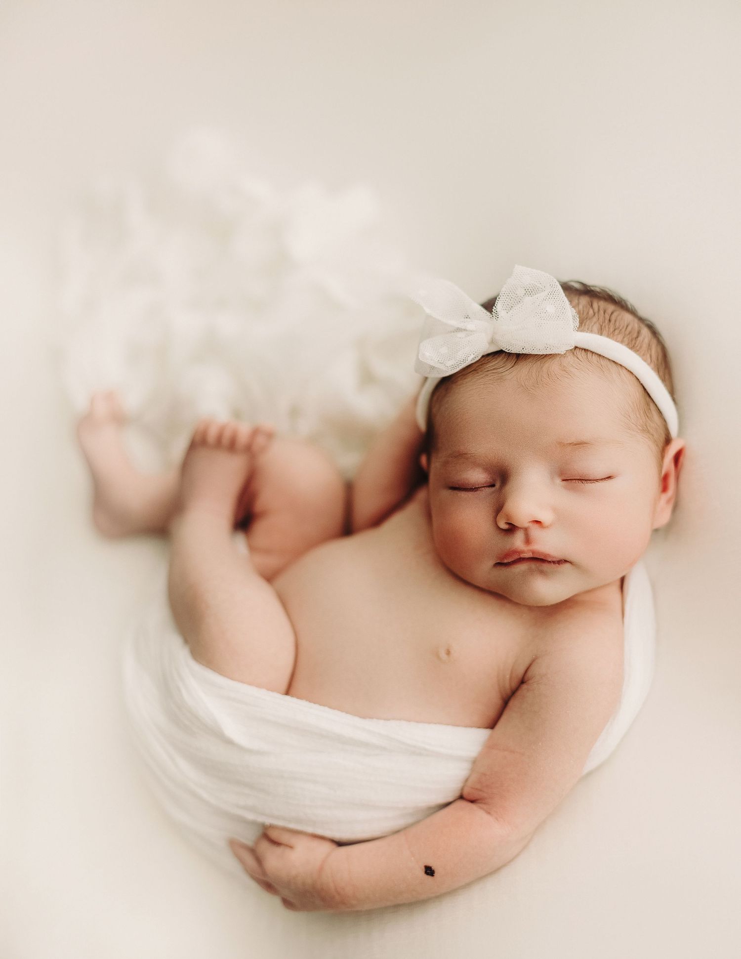 Peaceful sleeping newborn wrapped in soft white fabric wearing a delicate bow headband against cream background.