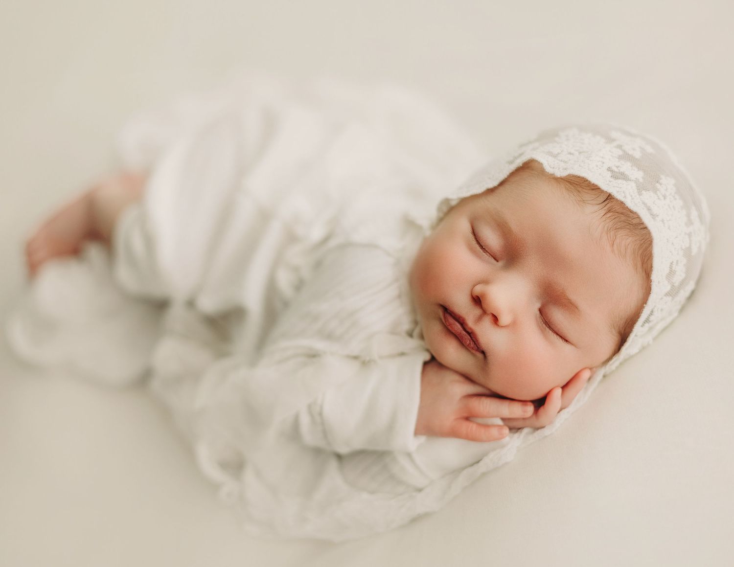 A newborn baby wearing a white lace bonnet sleeps peacefully on a soft neutral backdrop.