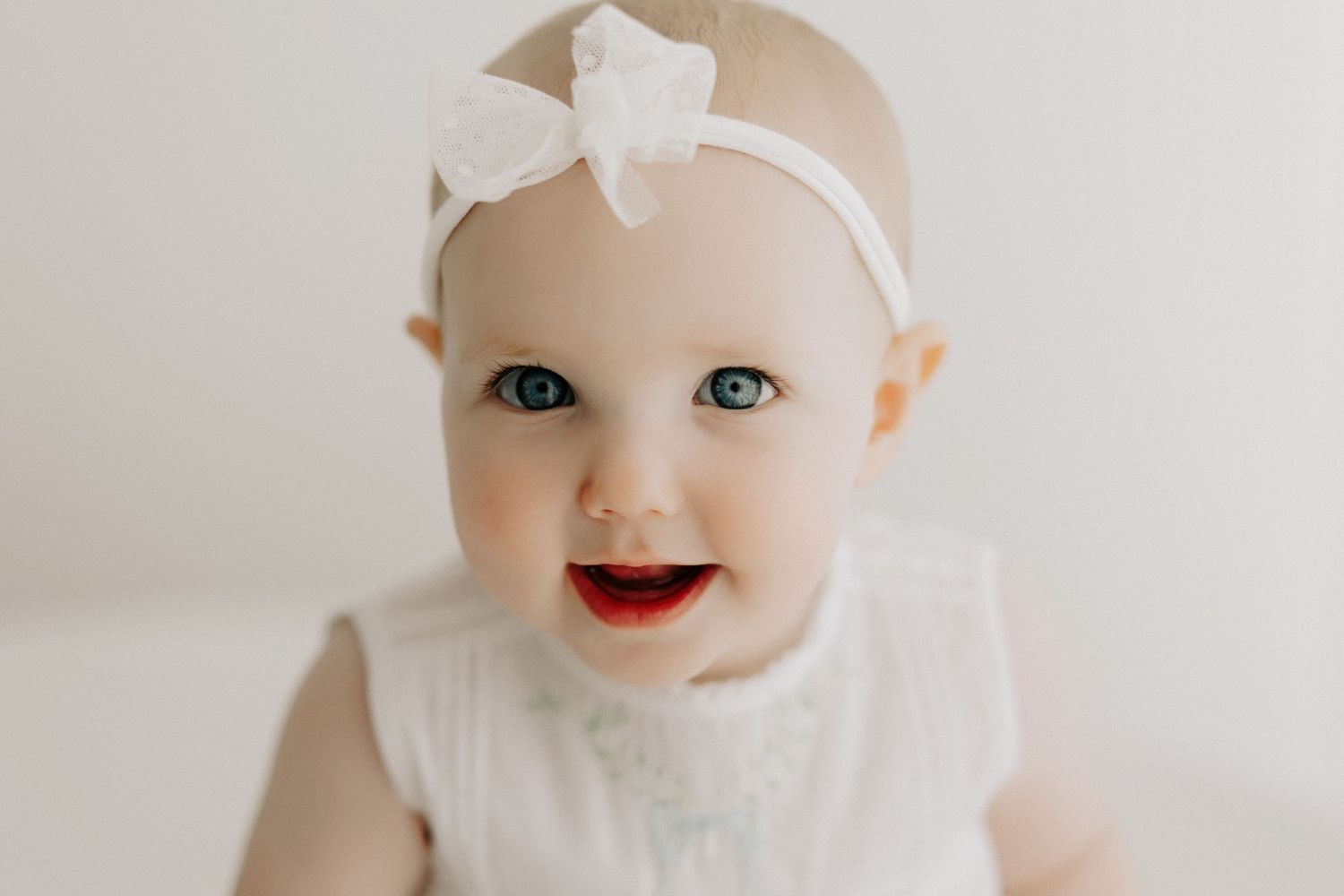 Smiling baby wearing white dress and bow headband against bright background.