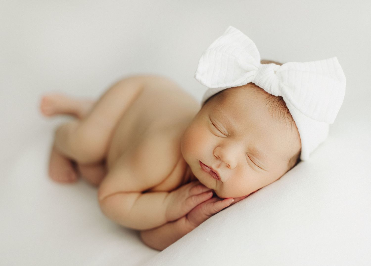 Sleeping newborn with white bow headband resting on light background.