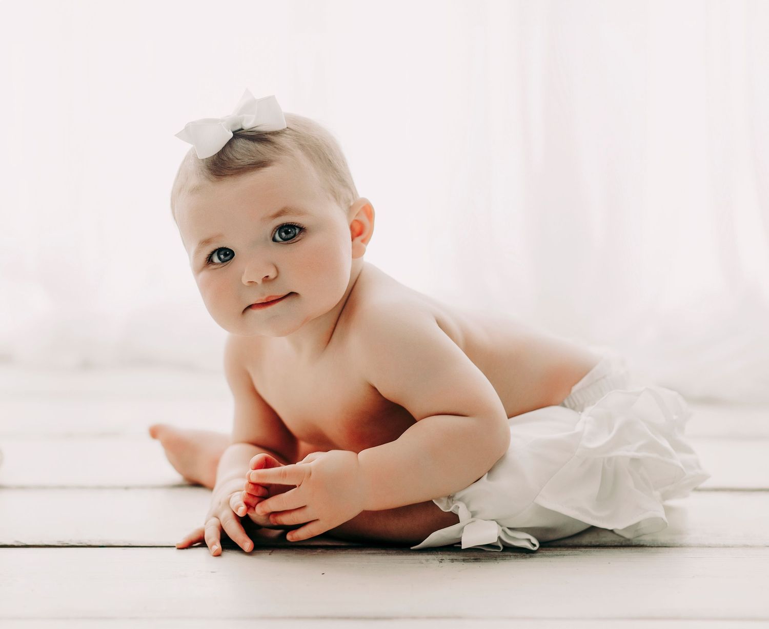 Baby in white diaper lying on white wooden floor with soft lighting.