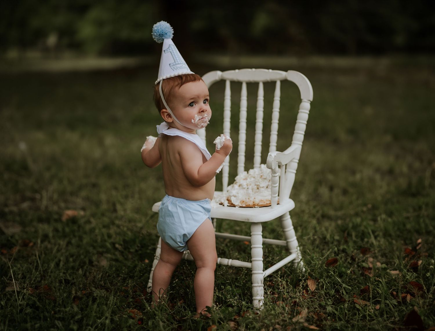 Baby in party hat standing by white chair during outdoor celebration.