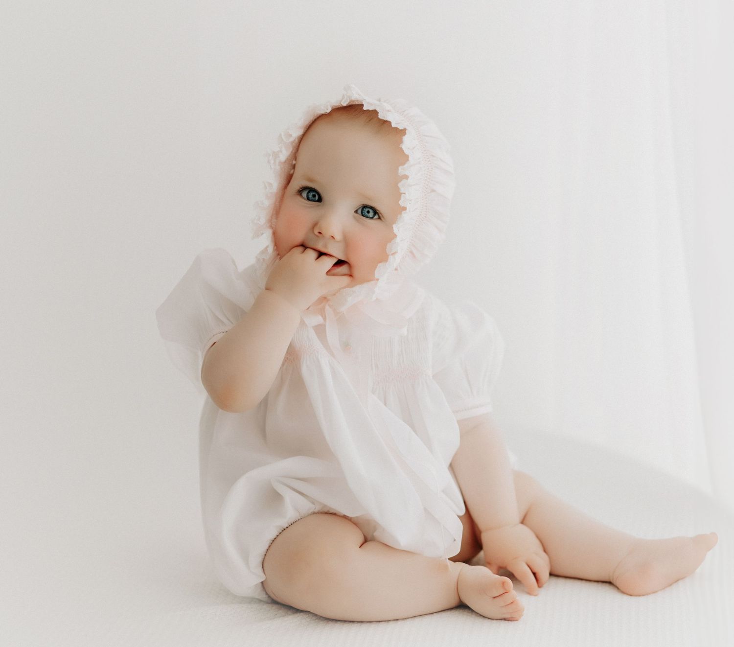 Baby in white lace outfit and bonnet sitting and chewing on fingers.