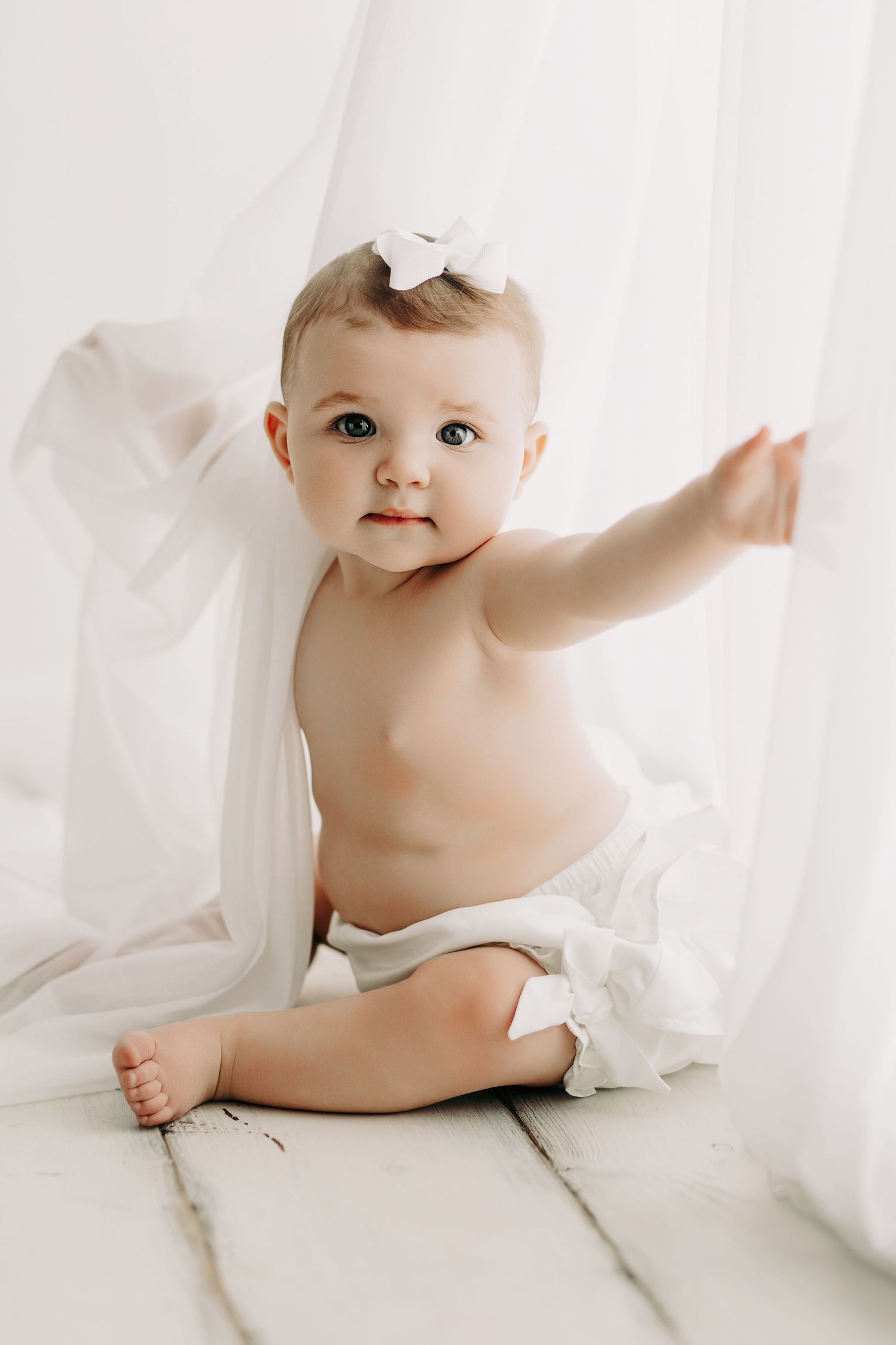 Happy baby wearing white bow sits on wooden floor with flowing white fabric.