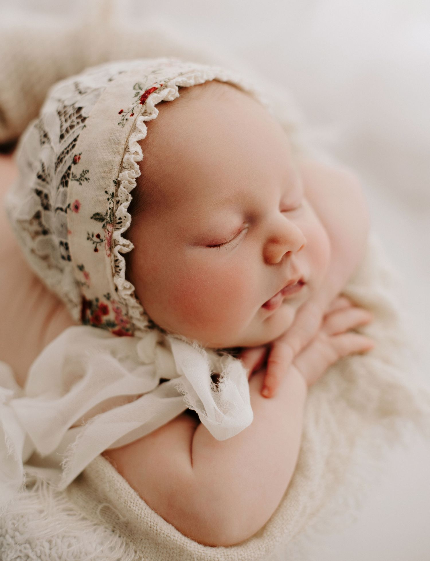 Sleeping newborn wearing floral bonnet rests peacefully on soft white blanket.