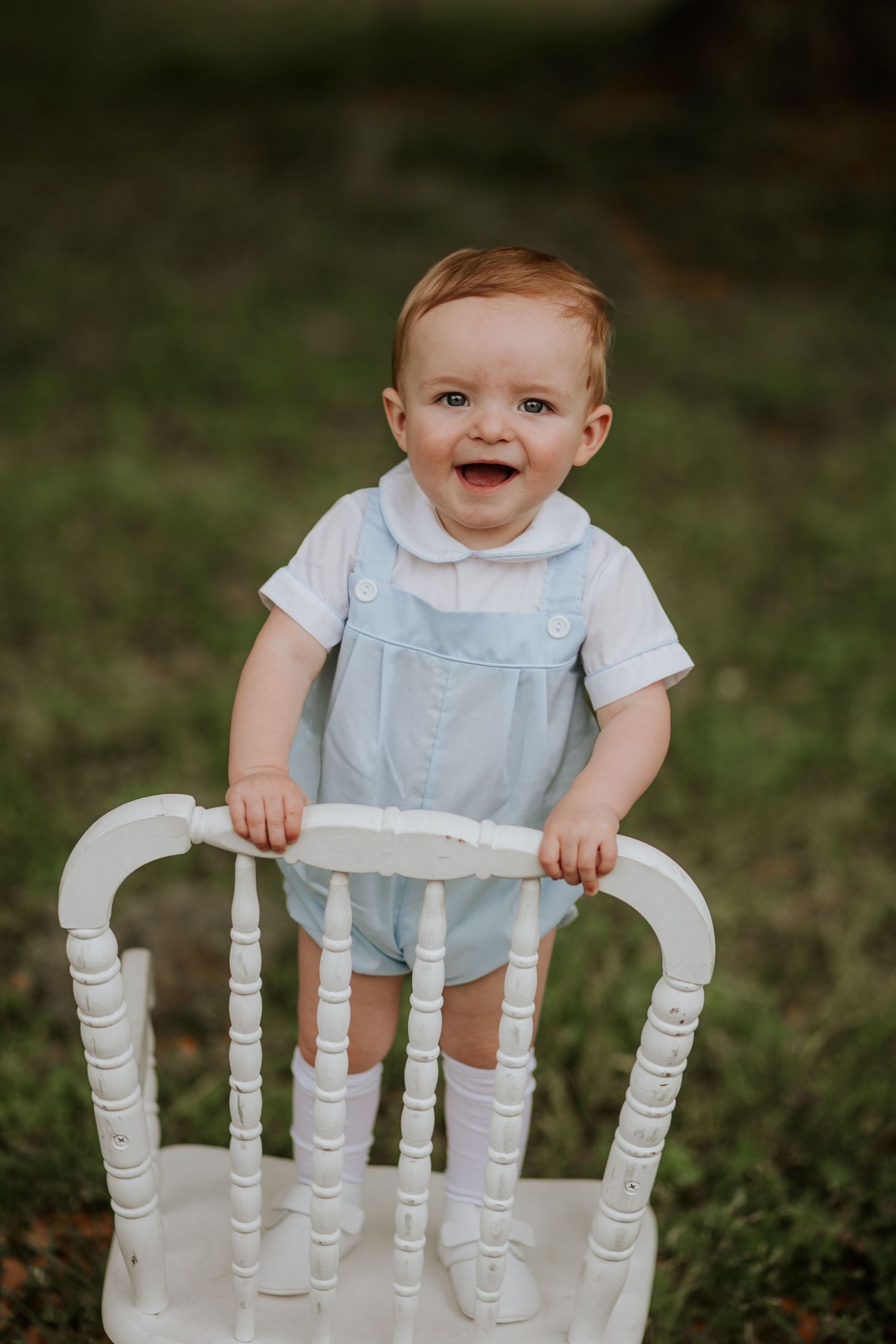 Toddler in blue outfit smiling while holding white vintage chair outdoors.