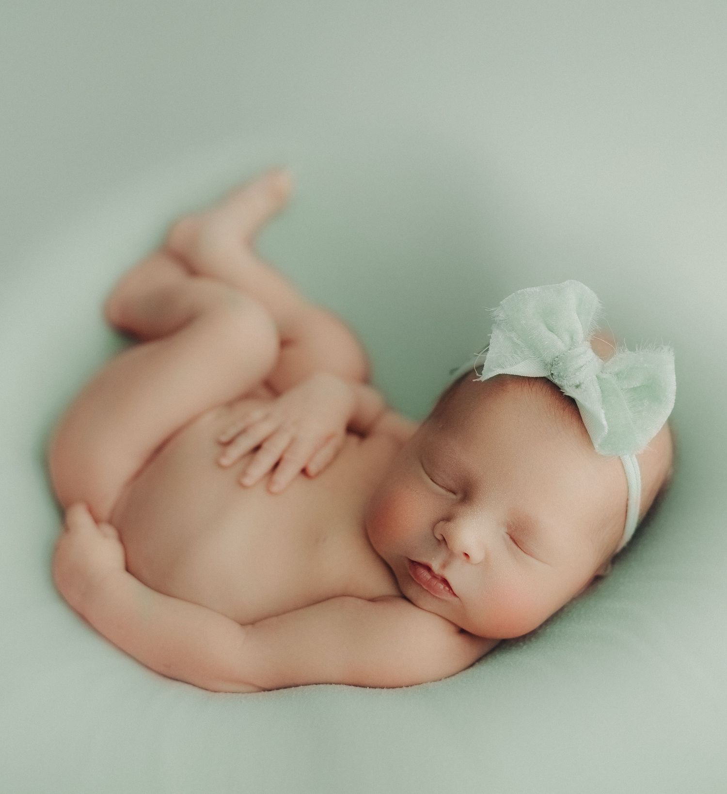 Peaceful sleeping newborn baby wearing white bow headband on mint green backdrop.