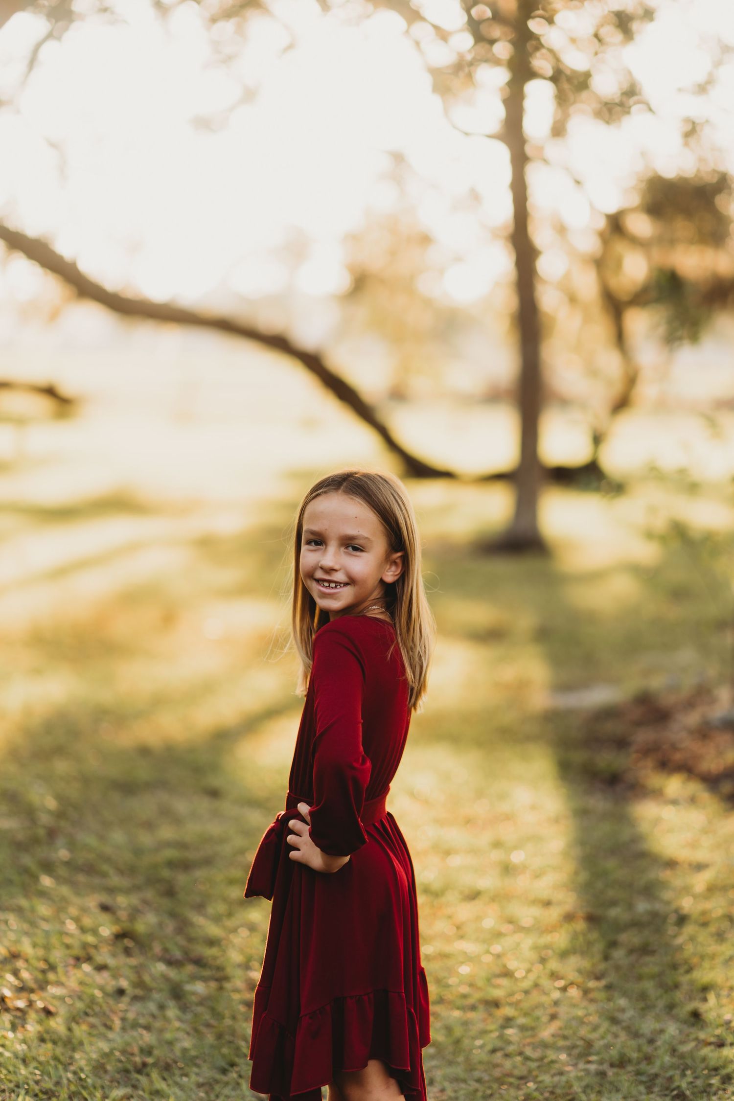Young child in red velvet dress twirls playfully in a sunny outdoor setting with trees in background.