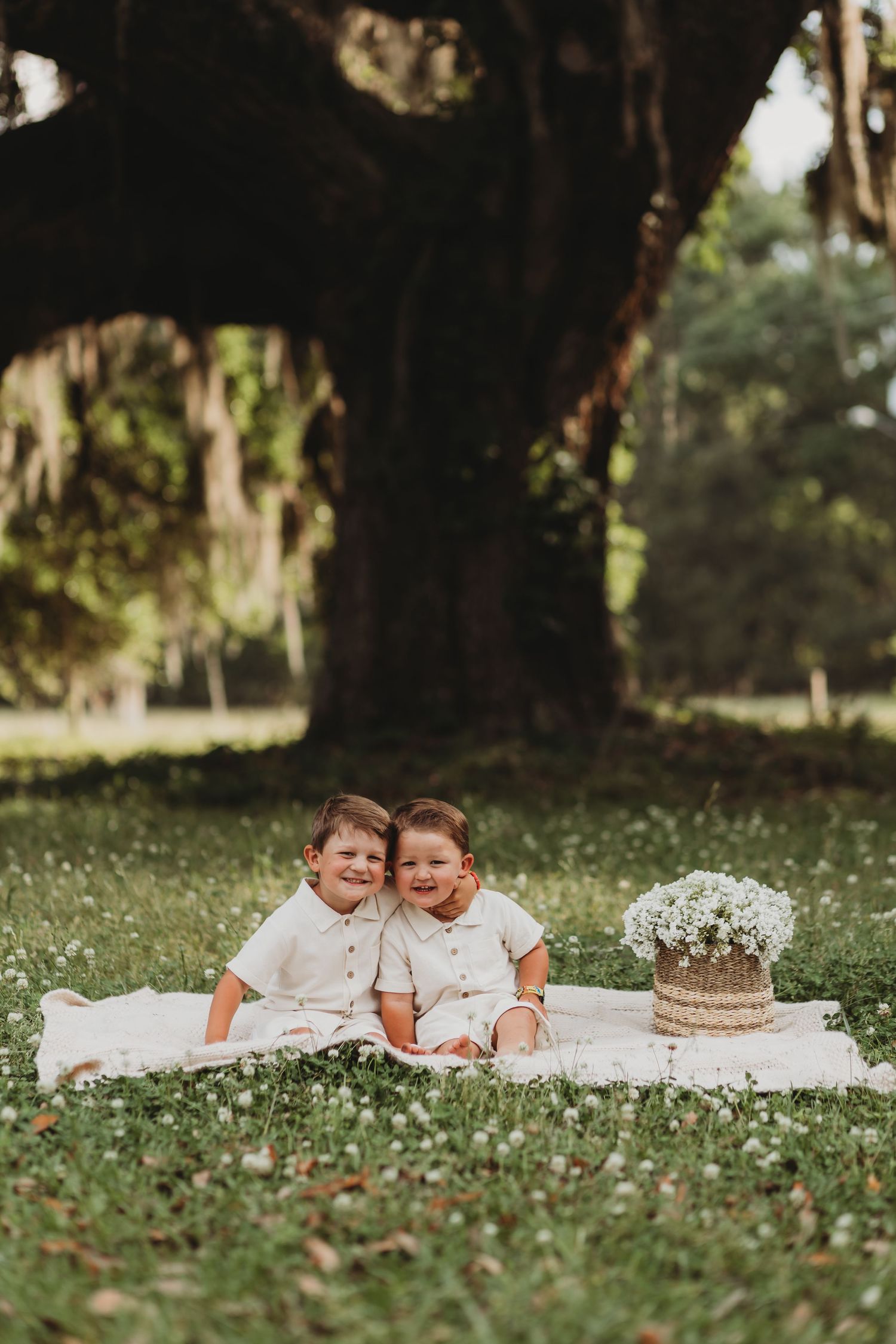 Two children lie on white blanket having picnic under large tree with flowers scattered around.
