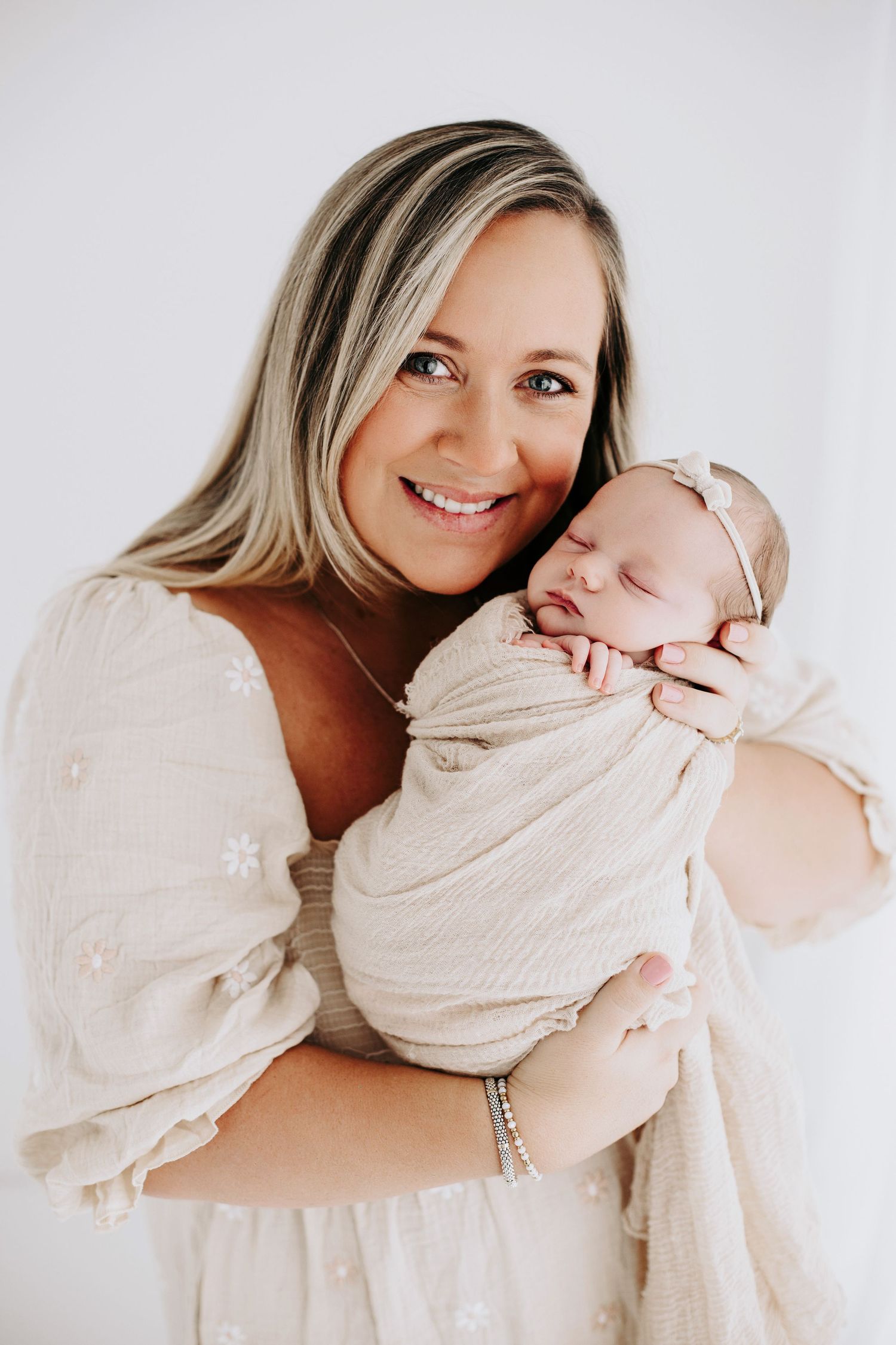 Mother in beige sweater lovingly embraces newborn baby in soft white outfit against light background.