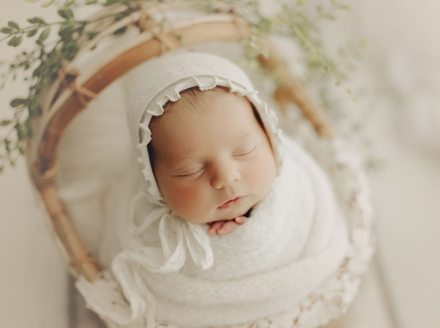 Newborn in white bonnet and wrap with delicate greenery decor.
