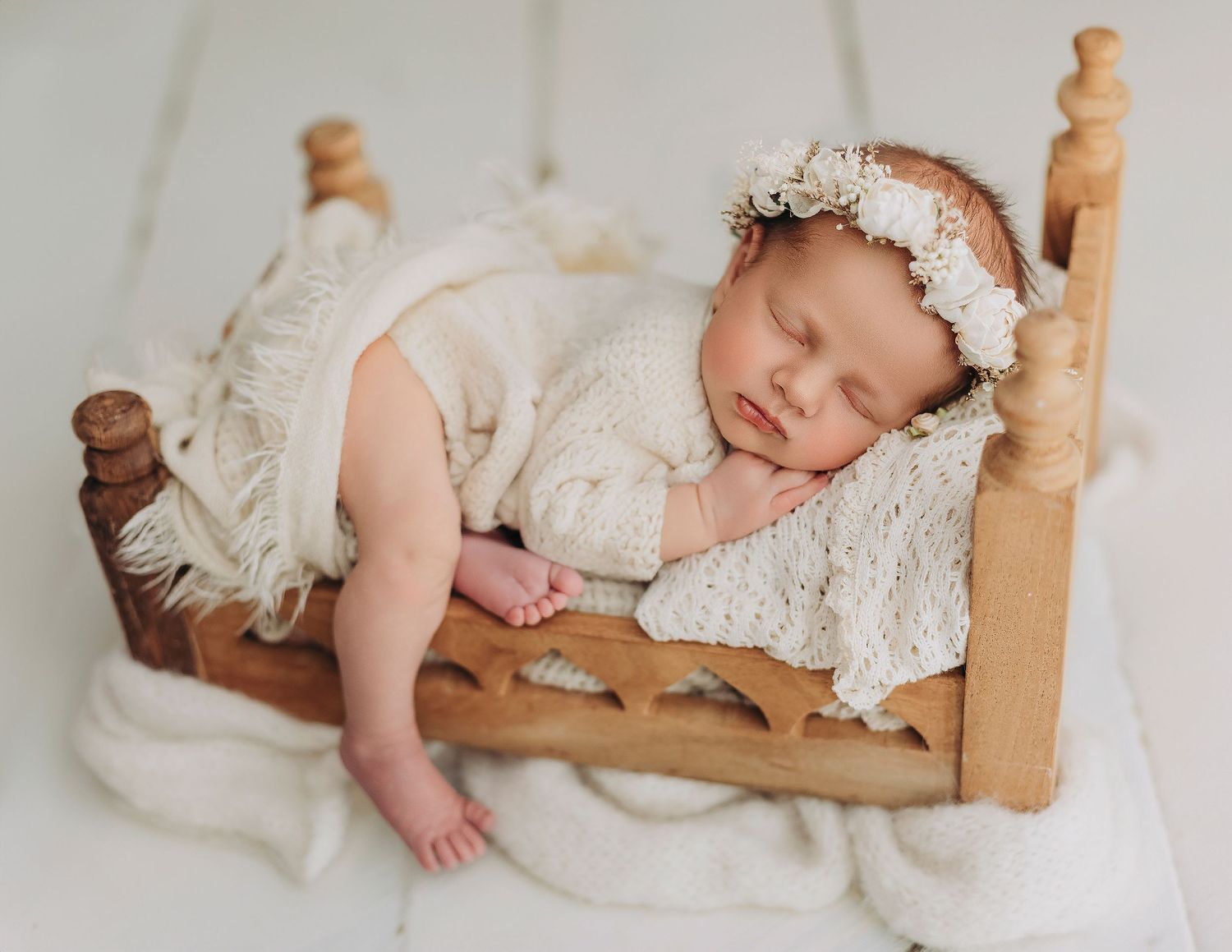 A sleeping baby wearing a white lace dress rests peacefully on a wooden bed with white bedding.