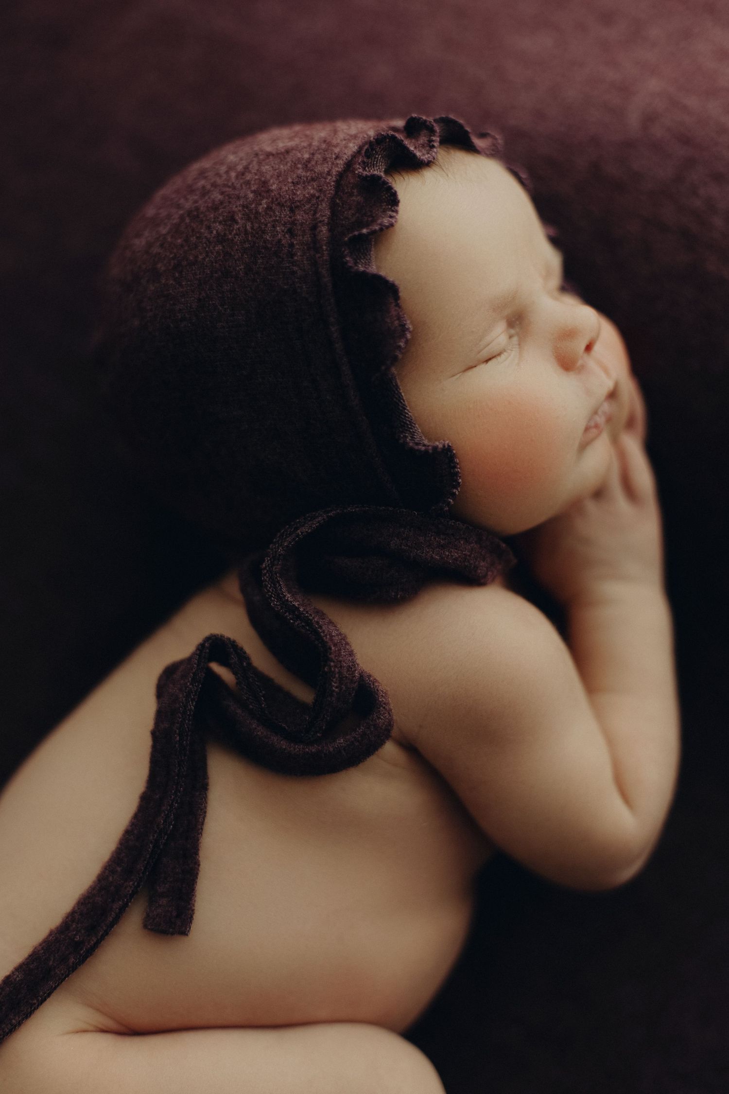 Peaceful newborn in brown bonnet resting hand near cheek on dark backdrop.