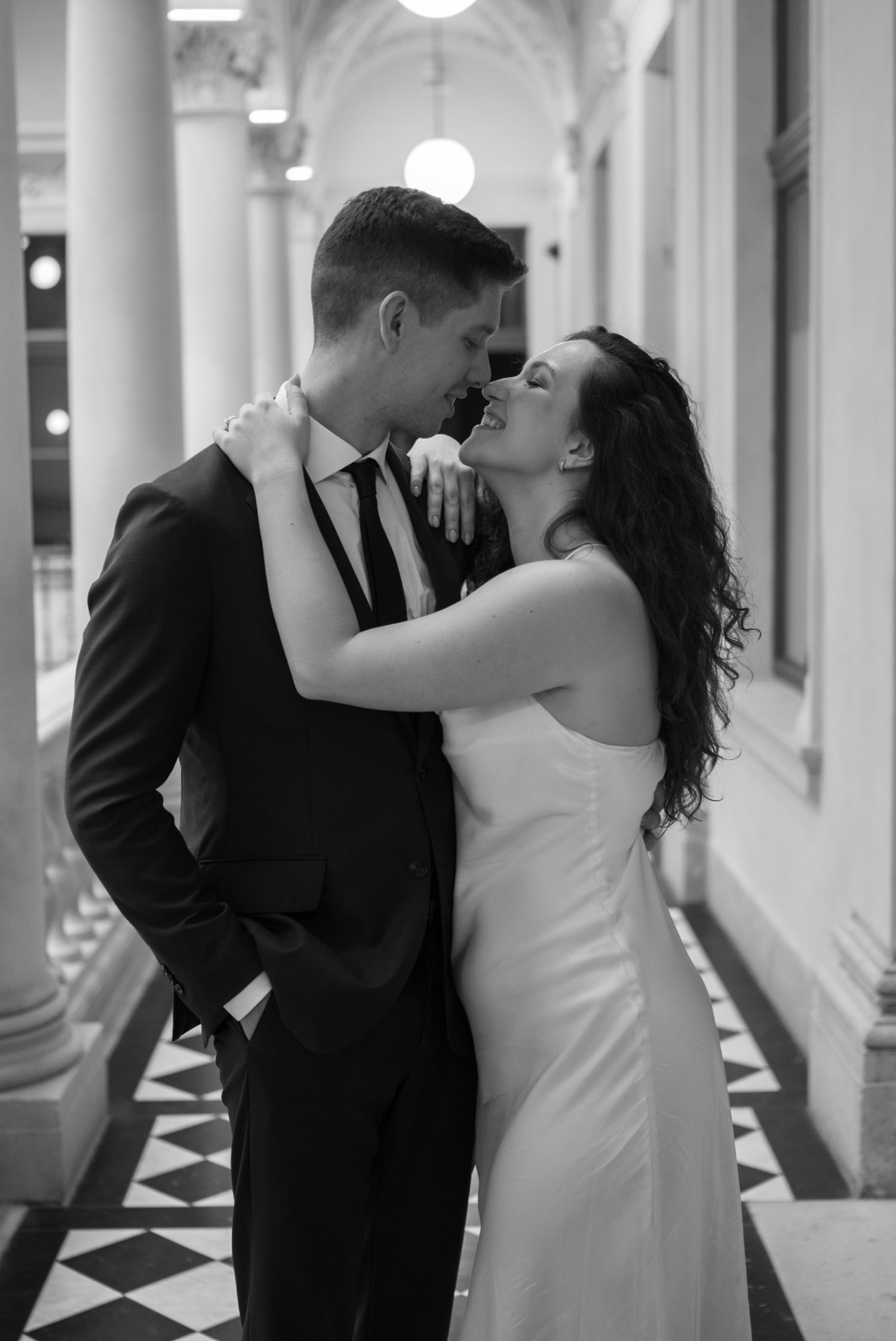 Black and white photo of a couple in wedding attire sharing a romantic moment in an elegant columned hallway.