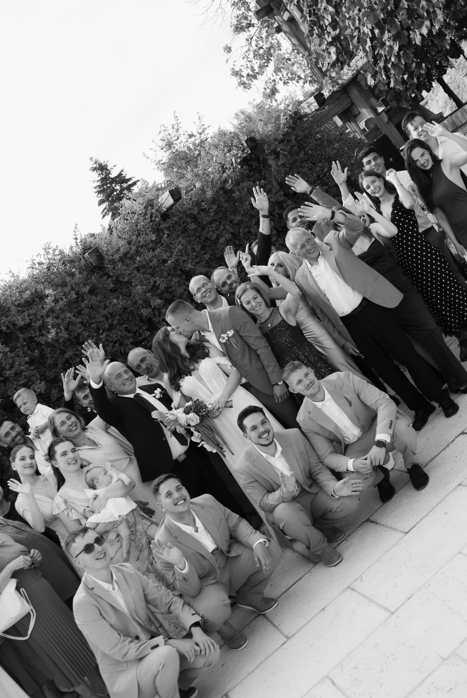 Black and white group photo of cheerful wedding guests waving at outdoor summer wedding celebration.