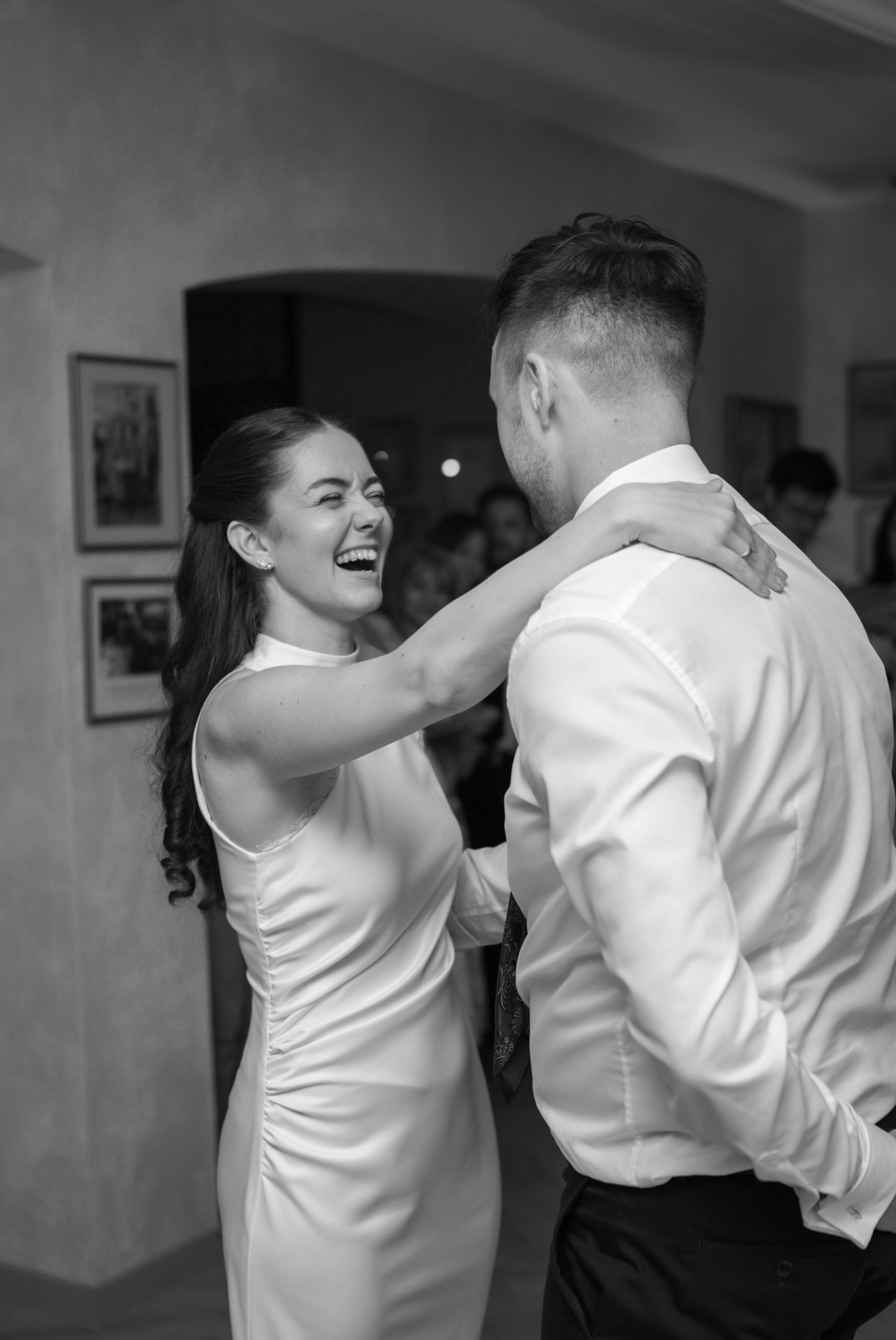 Bride in white halter dress laughs joyfully while sharing first dance with groom in black and white photo.