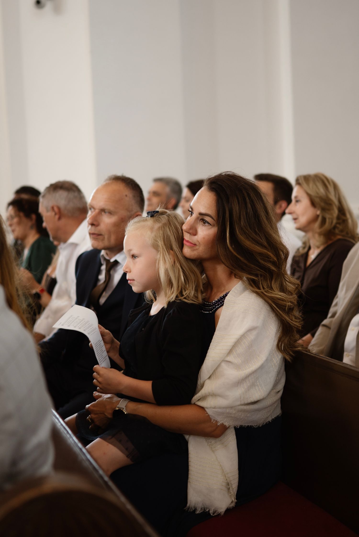 A woman with long brown hair holds a young blonde girl on her lap during a church wedding ceremony.