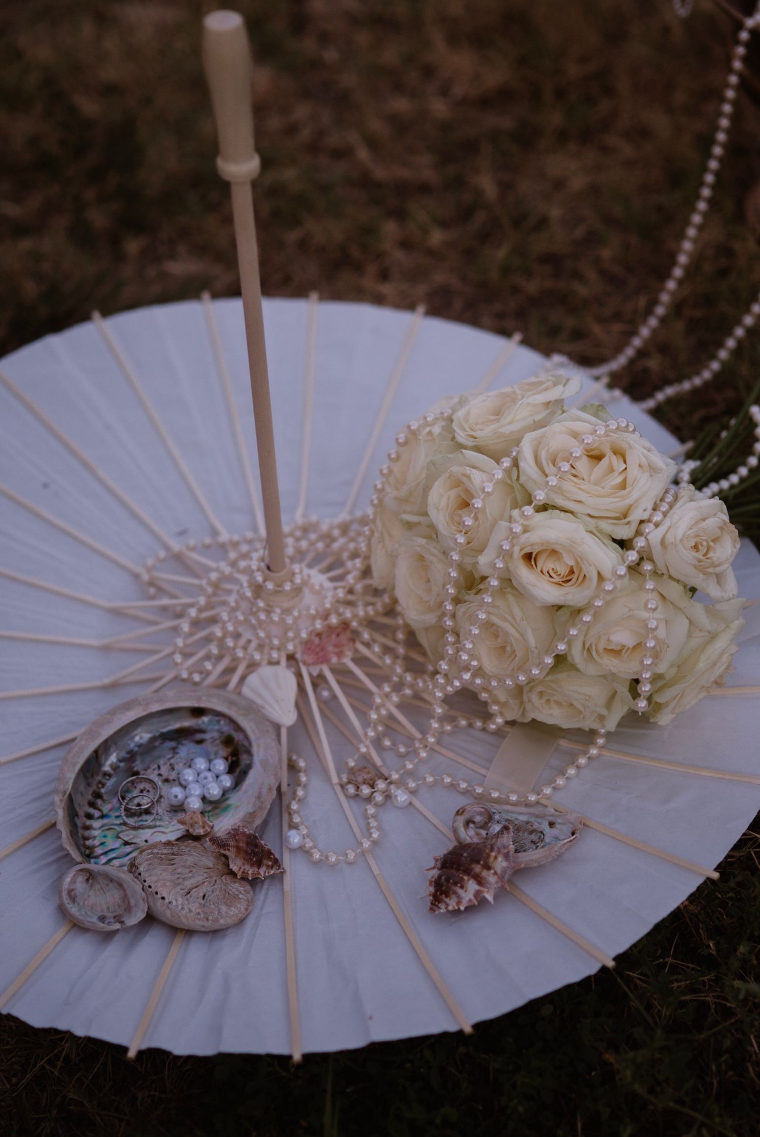 White parasol with cream roses bouquet, pearl necklace, and seashells arranged elegantly on grass.