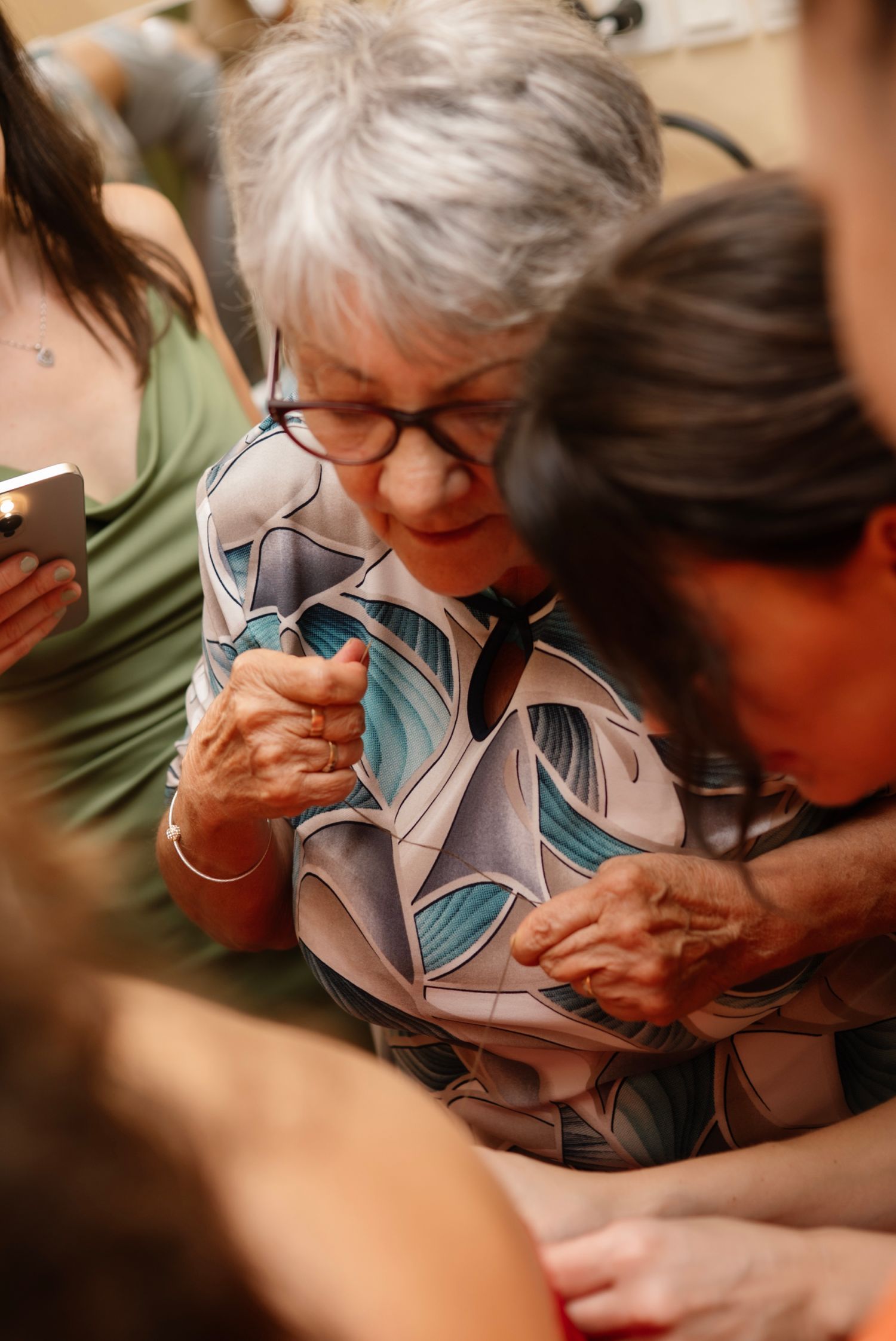 Elderly woman with gray hair and glasses examines something closely, wearing a colorful patterned blouse.