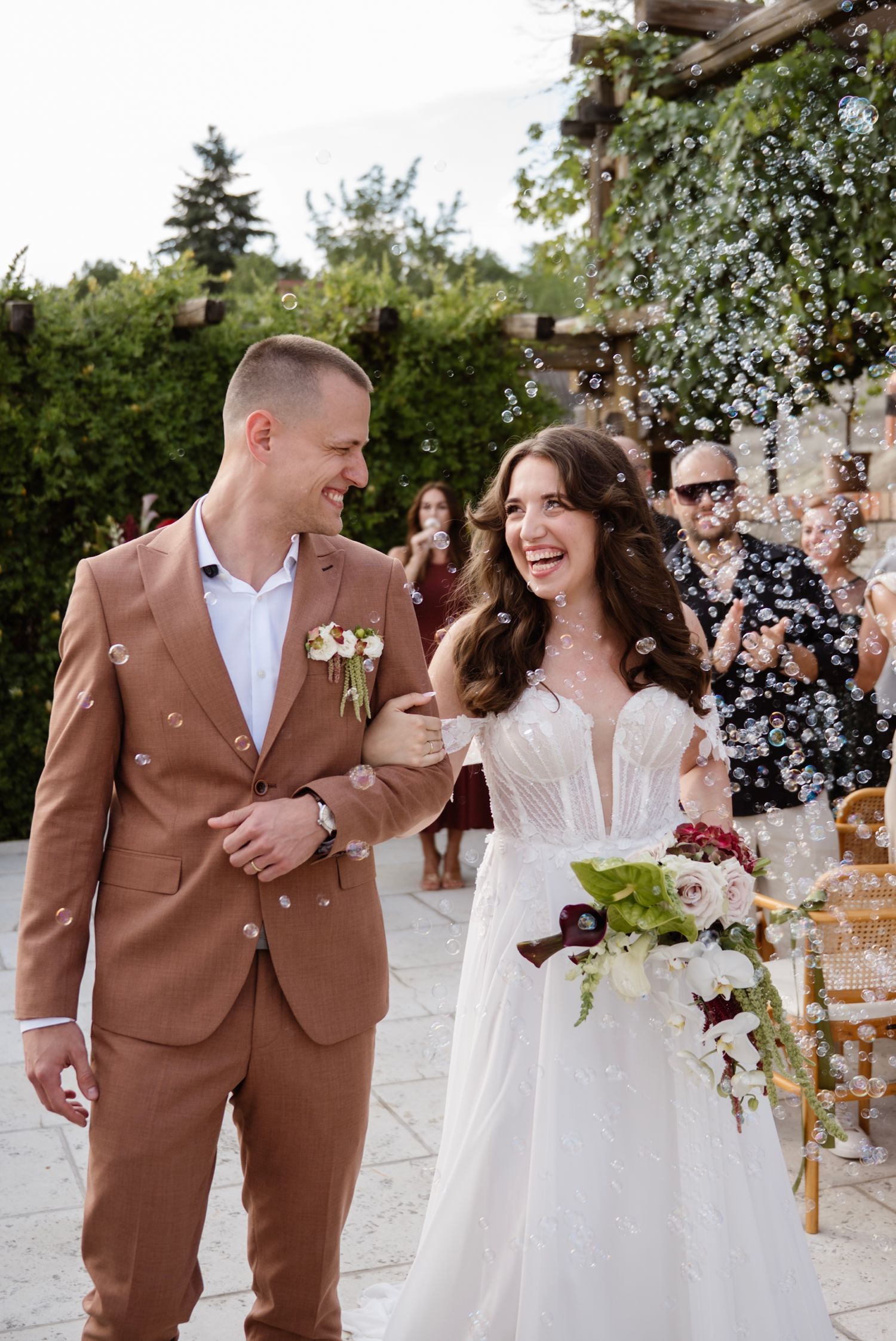 Bride and groom walk joyfully through bubble send-off at outdoor wedding ceremony with guests cheering.