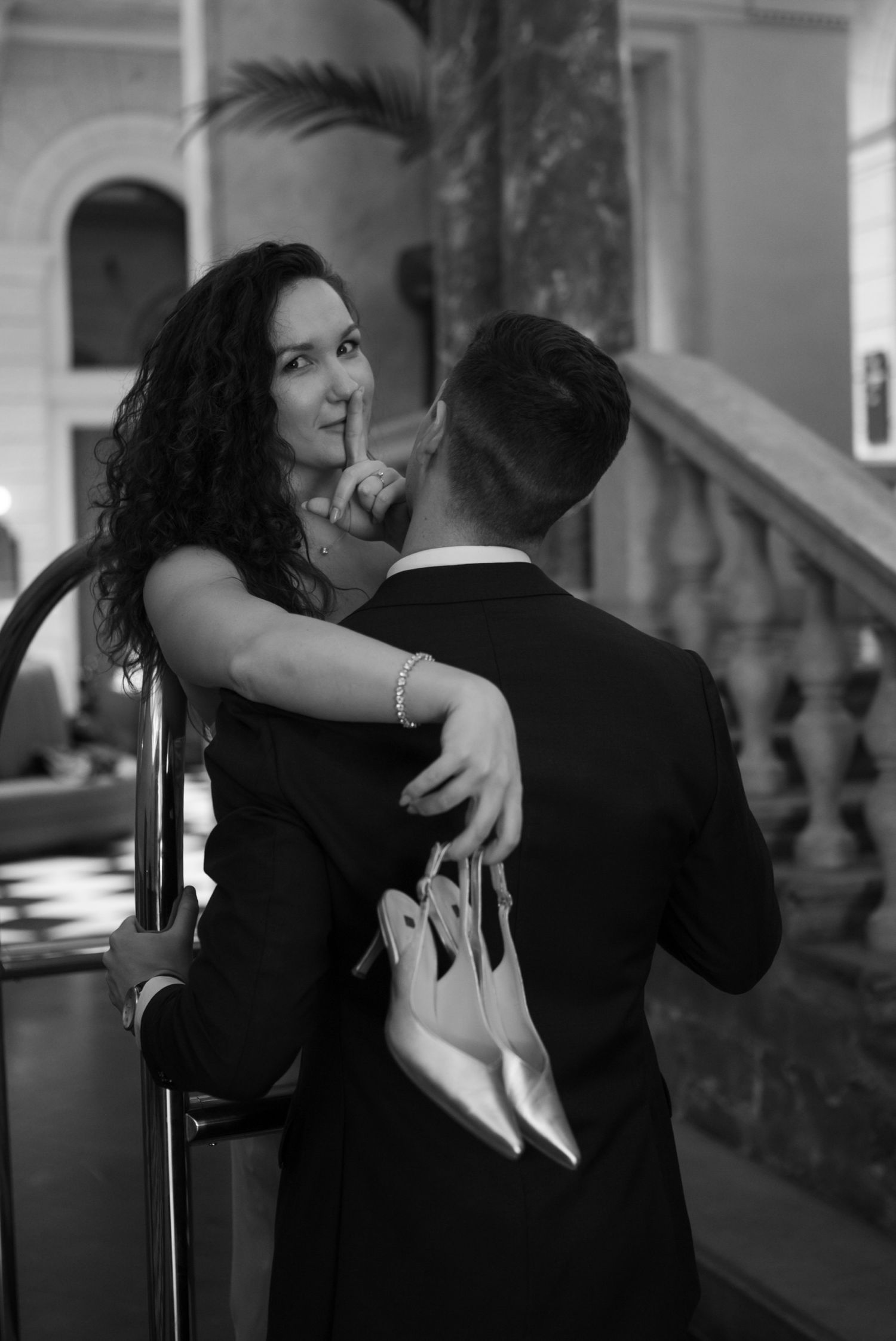 Black and white photo of a couple embracing near a staircase, woman holding heels and gesturing silence.