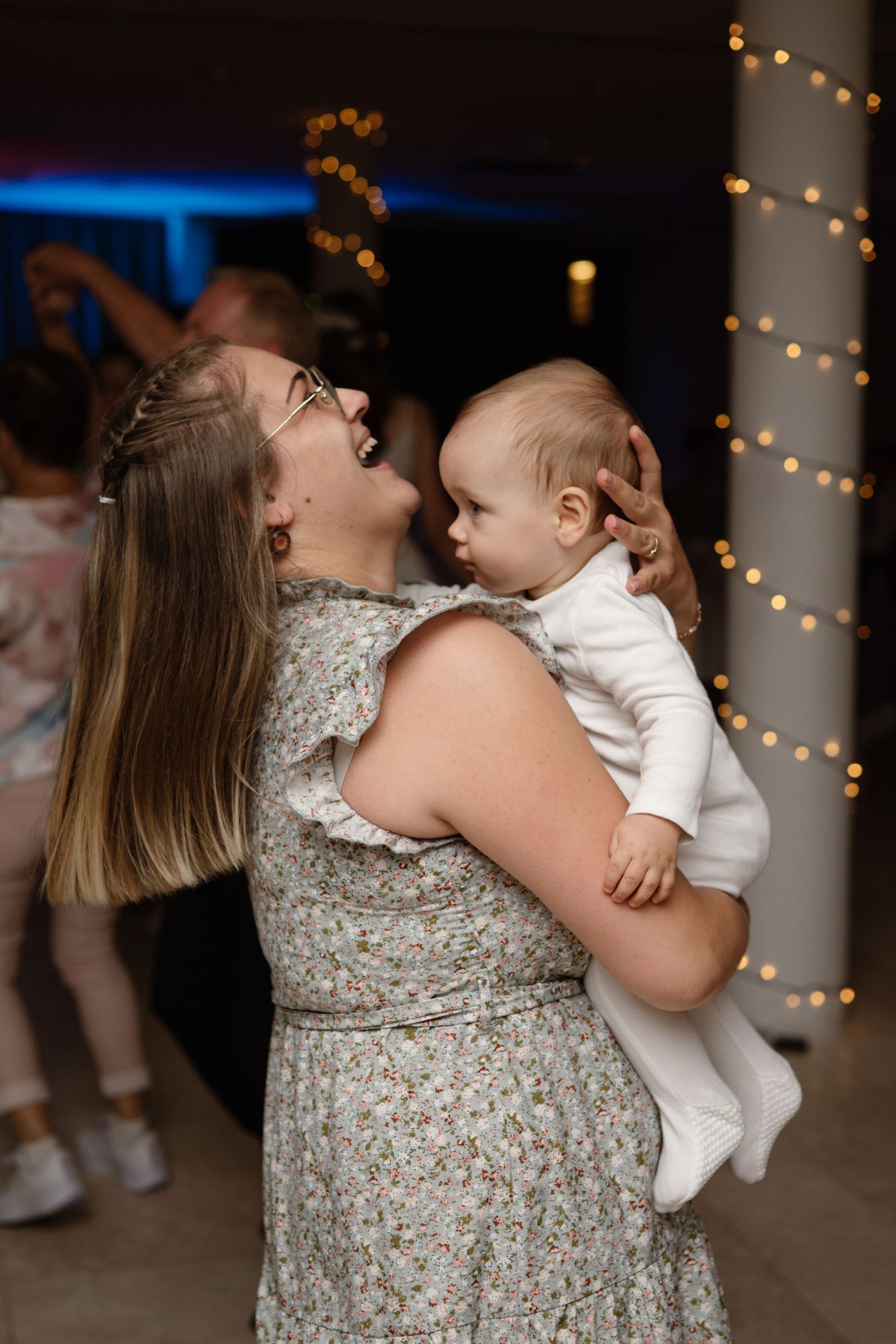 A woman in a floral dress dances with a baby at a wedding reception with string lights in the background.