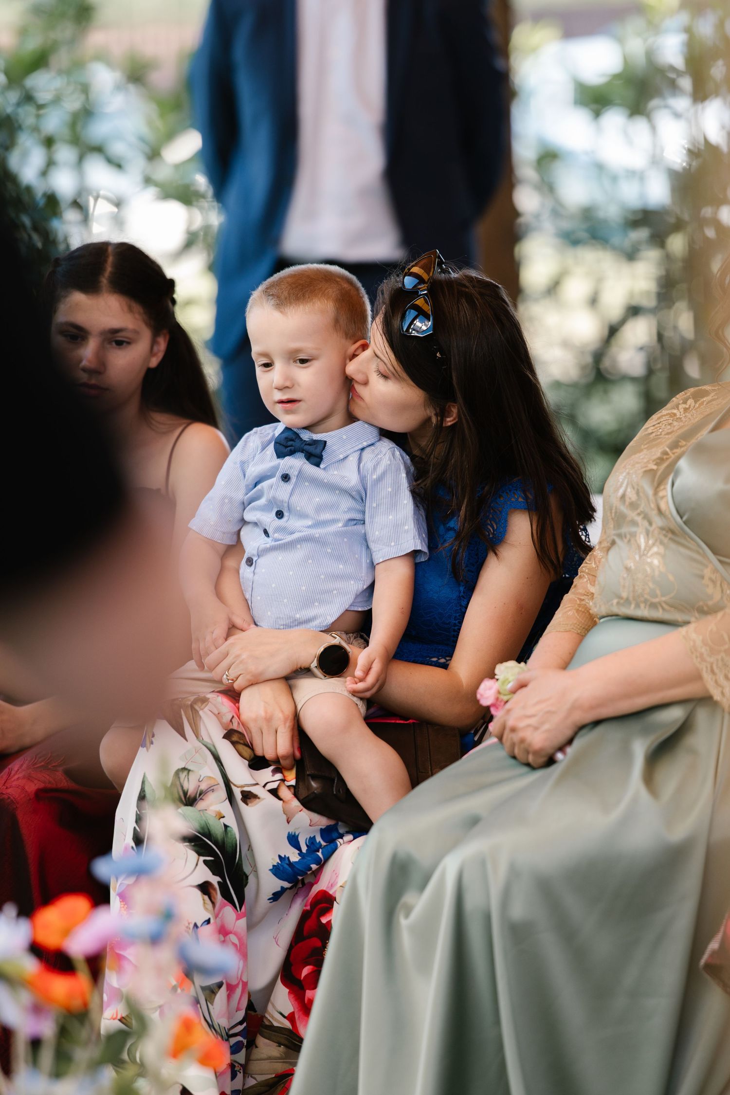 Young child wearing sunglasses makes silly face while sitting with adults at outdoor event.