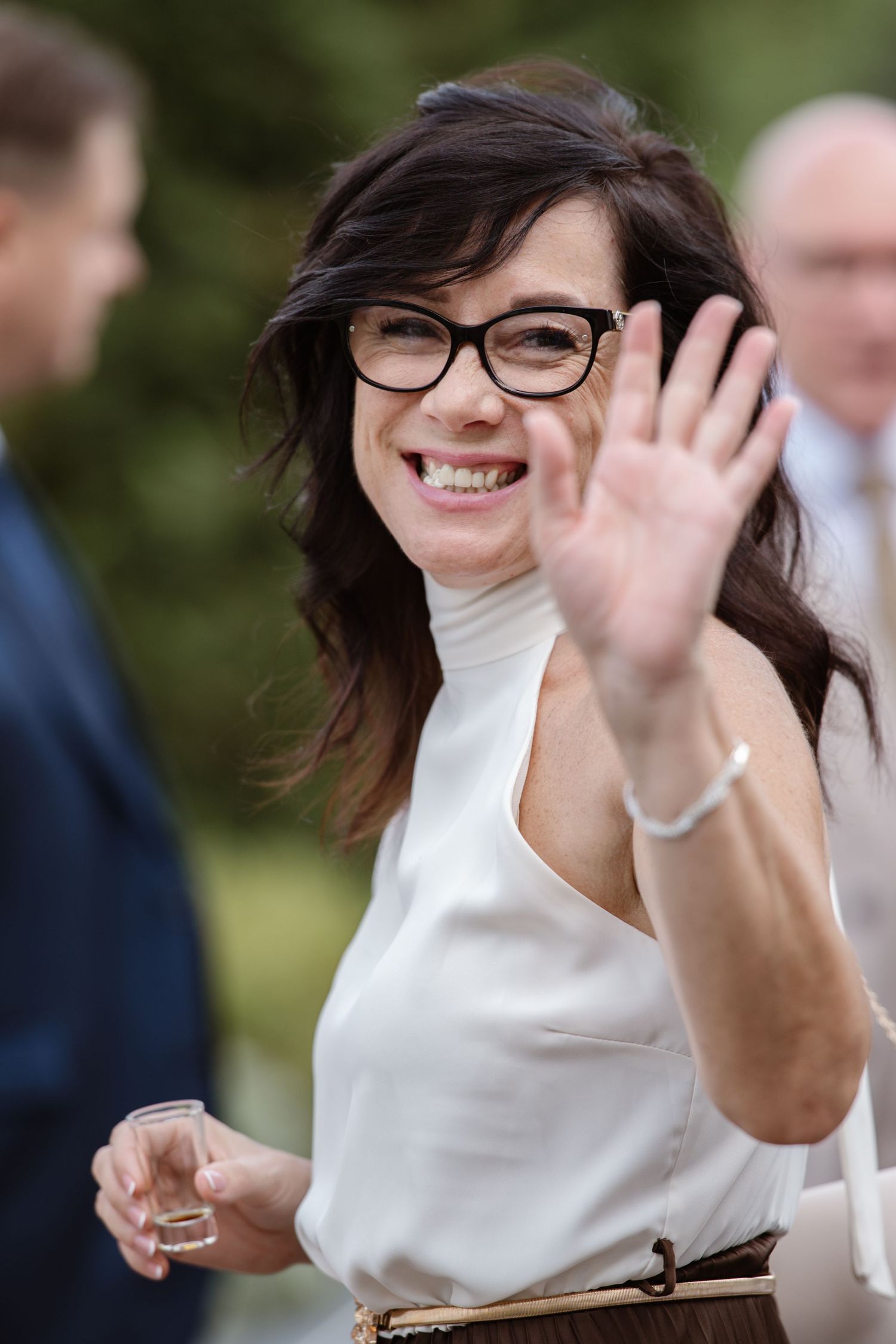 A person in a white top and glasses waving enthusiastically at an outdoor gathering.