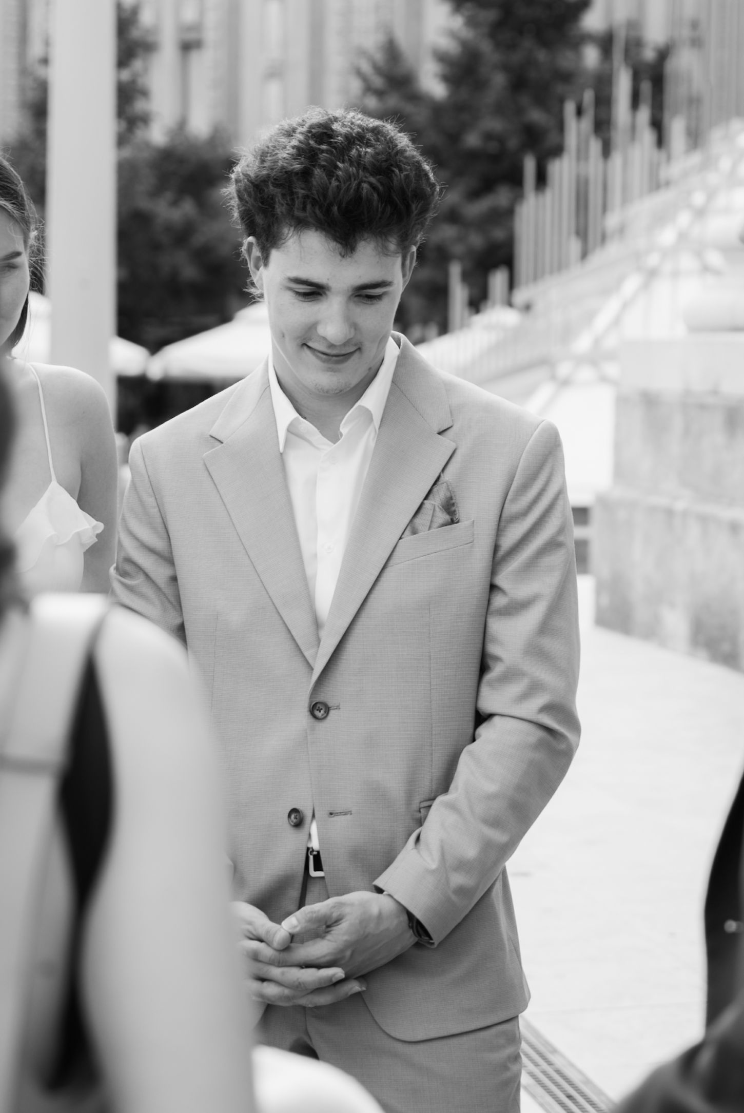 Young man in light suit smiling with clasped hands at outdoor wedding ceremony, black and white photo.