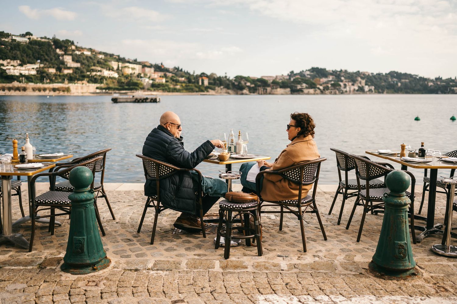 People enjoying drinks at outdoor waterfront cafe tables with scenic coastal views of a Mediterranean harbor at sunset.