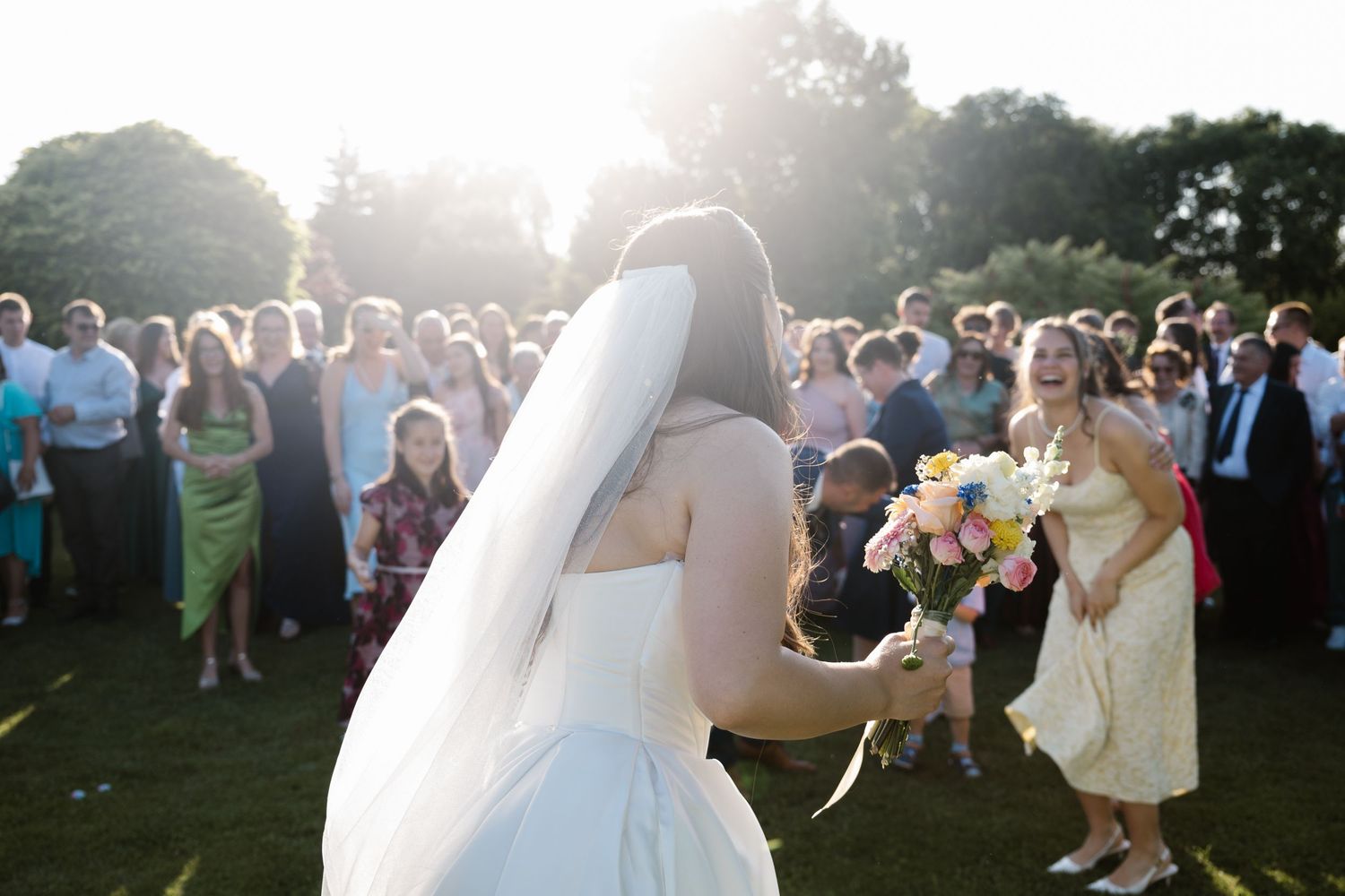 A bride prepares to toss her bouquet to a crowd of wedding guests gathered on a sunny lawn.